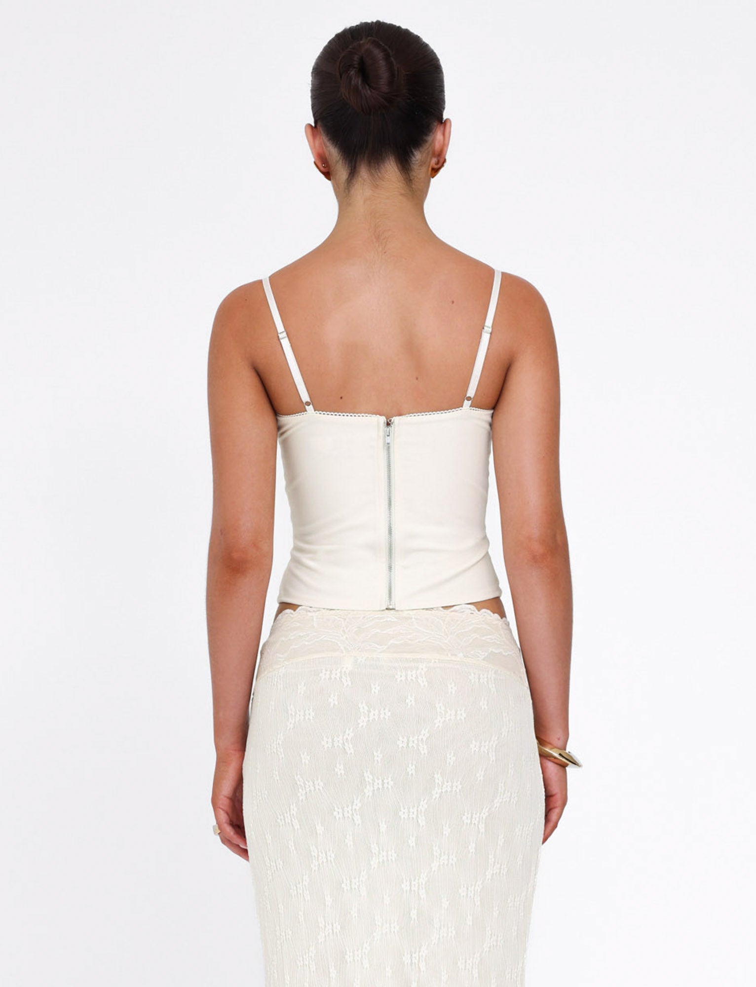 A woman faces away from the camera in the TIGERMIST ANTONELLA TOP—bone cream color with adjustable straps and a visible back zipper—paired with a sheer lace-patterned skirt; her dark hair is styled in a neat bun.