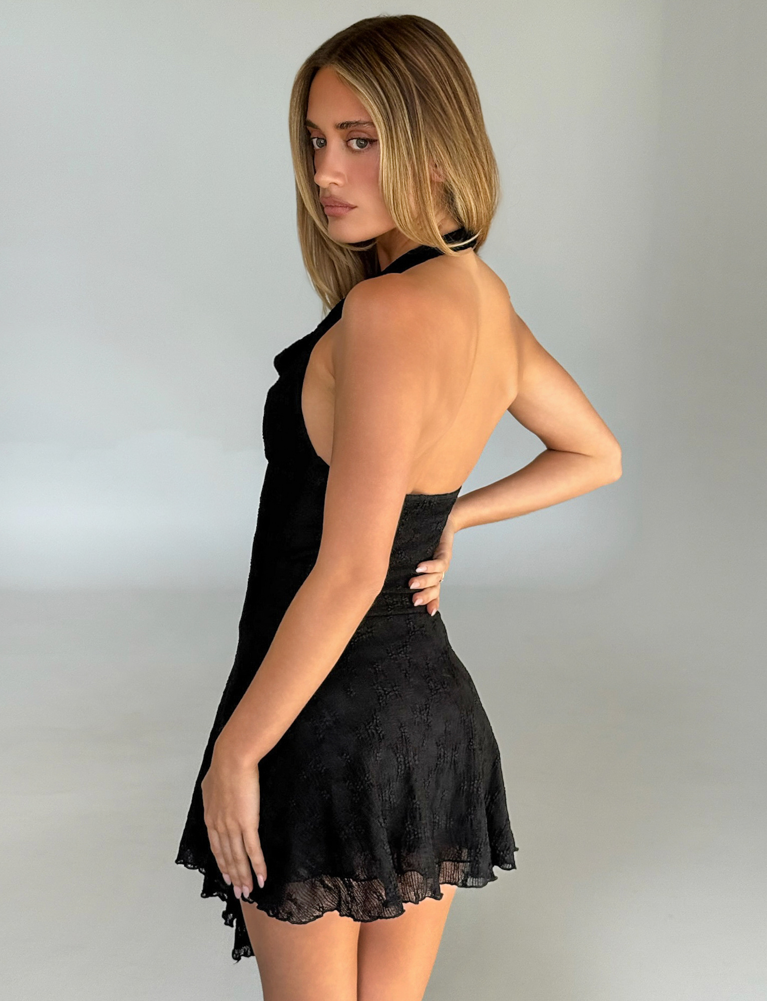 A blonde woman poses sideways, looking over her shoulder, wearing the TIGERMIST FIORELLA DRESS in black lace— a halter mini dress with a fit and flare silhouette—against a plain, light background.