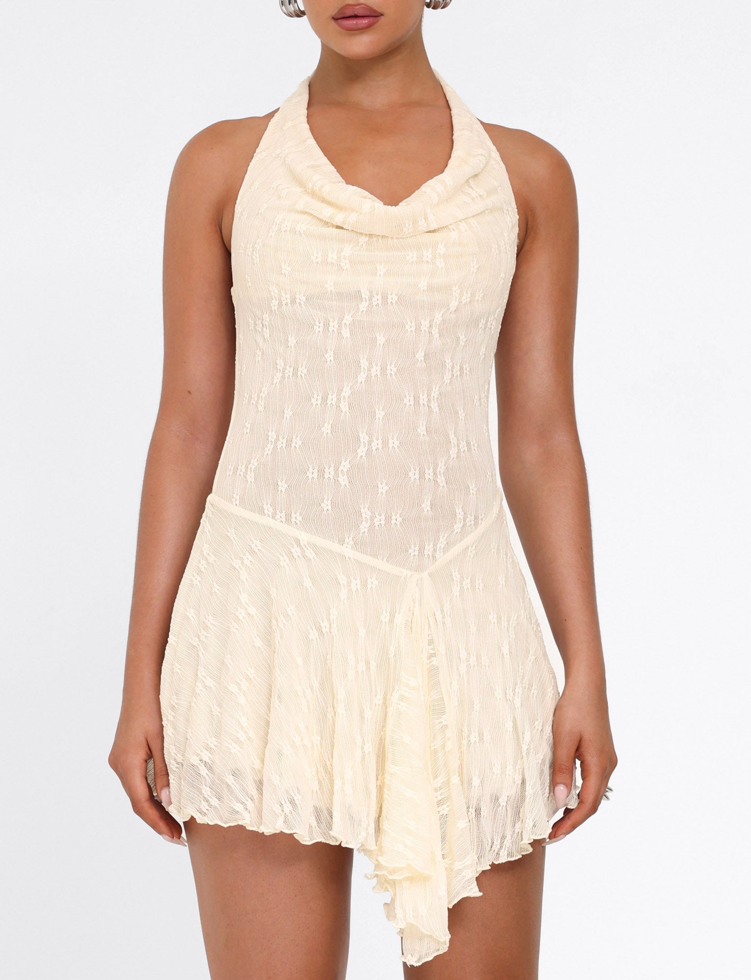 A woman models the TIGERMIST FIORELLA MINI DRESS—sleeveless, cream lace with a halter neck, cowl neckline, and asymmetrical hem—against a plain white background.