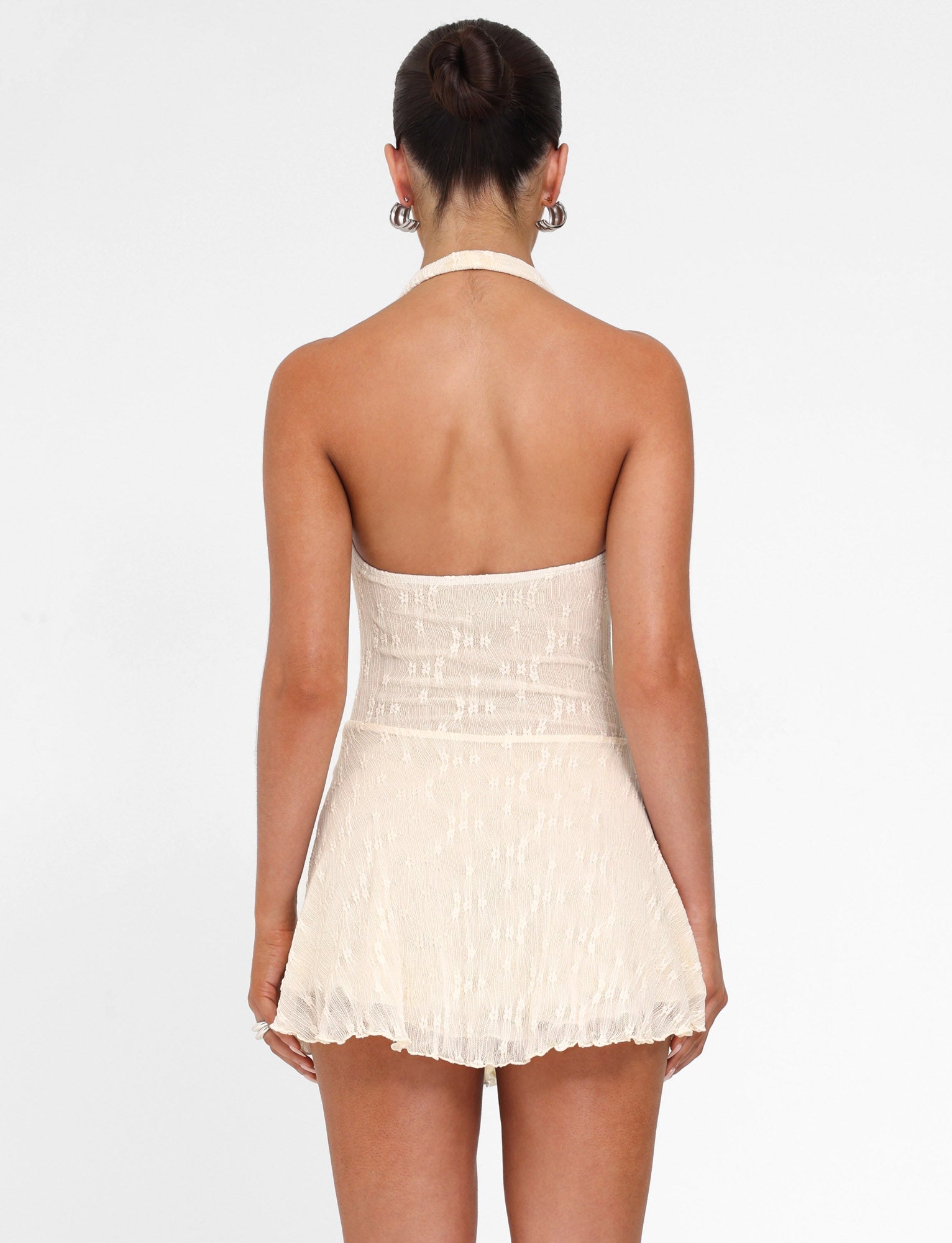 A woman with a neat bun faces away from the camera, wearing the TIGERMIST FIORELLA MINI DRESS in cream lace. The stylish dress features a halter neck and fitted waist against a plain white background.