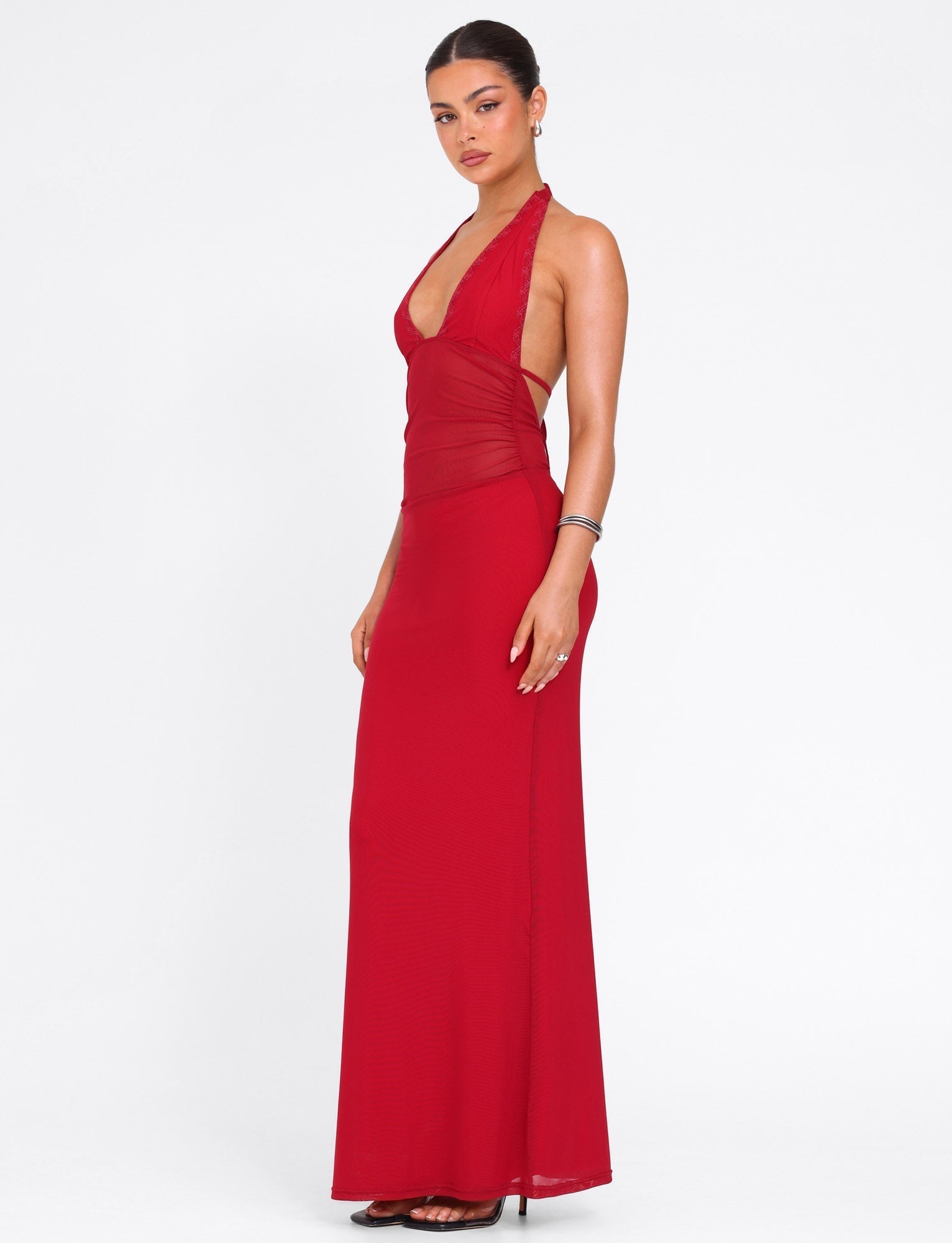A woman stands in profile against a white background wearing the TIGERMIST BRESILY MAXI DRESS in red, a sleeveless floor-length gown with halter straps and a fitted bodice, styled with black heels and minimal jewelry. Her hair is pulled back.