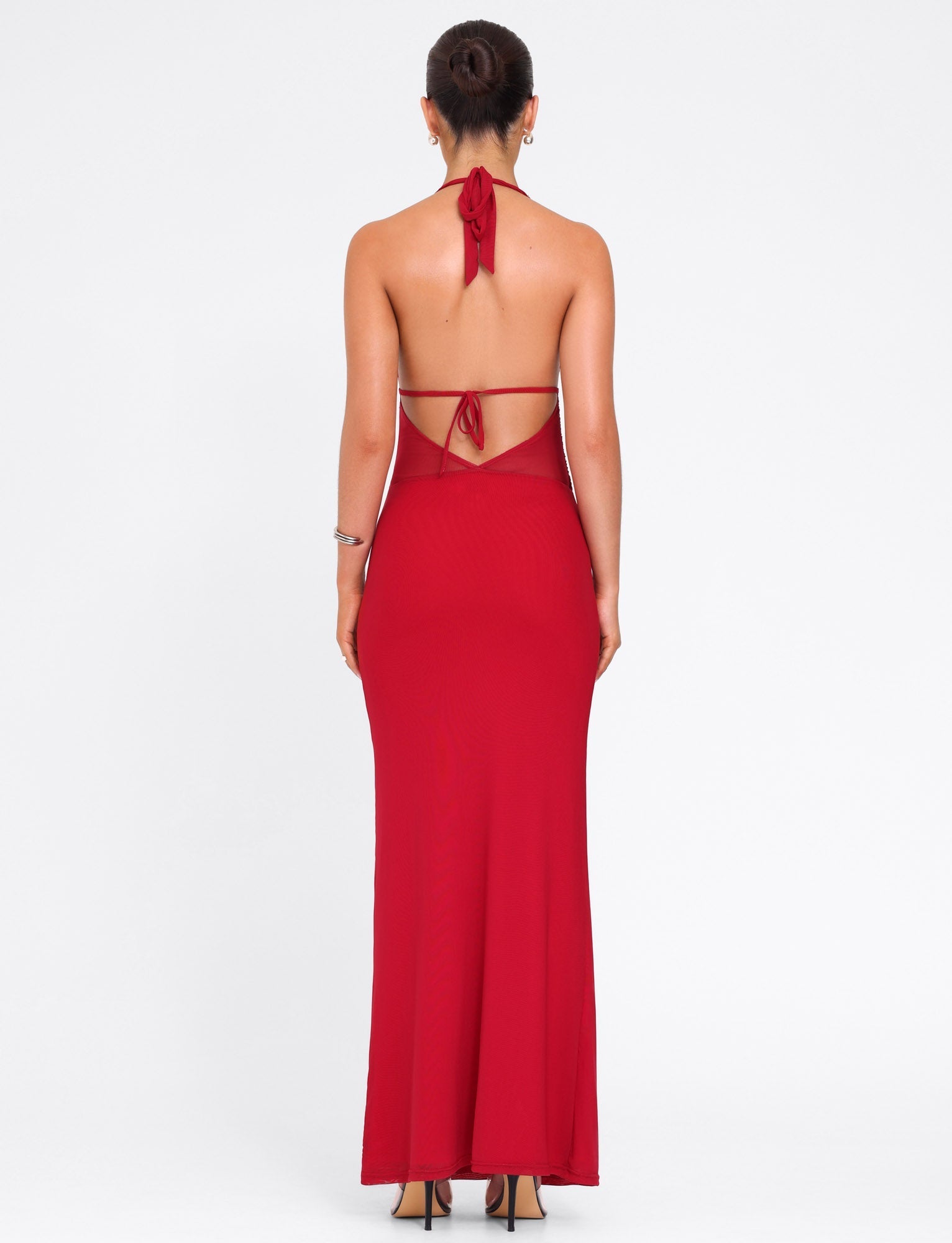 A woman with her hair up wears the TIGERMIST BRESILY MAXI DRESS in red—a long, backless halter style tied at the neck and mid-back. She pairs it with high heels and hoop earrings against a plain white background.