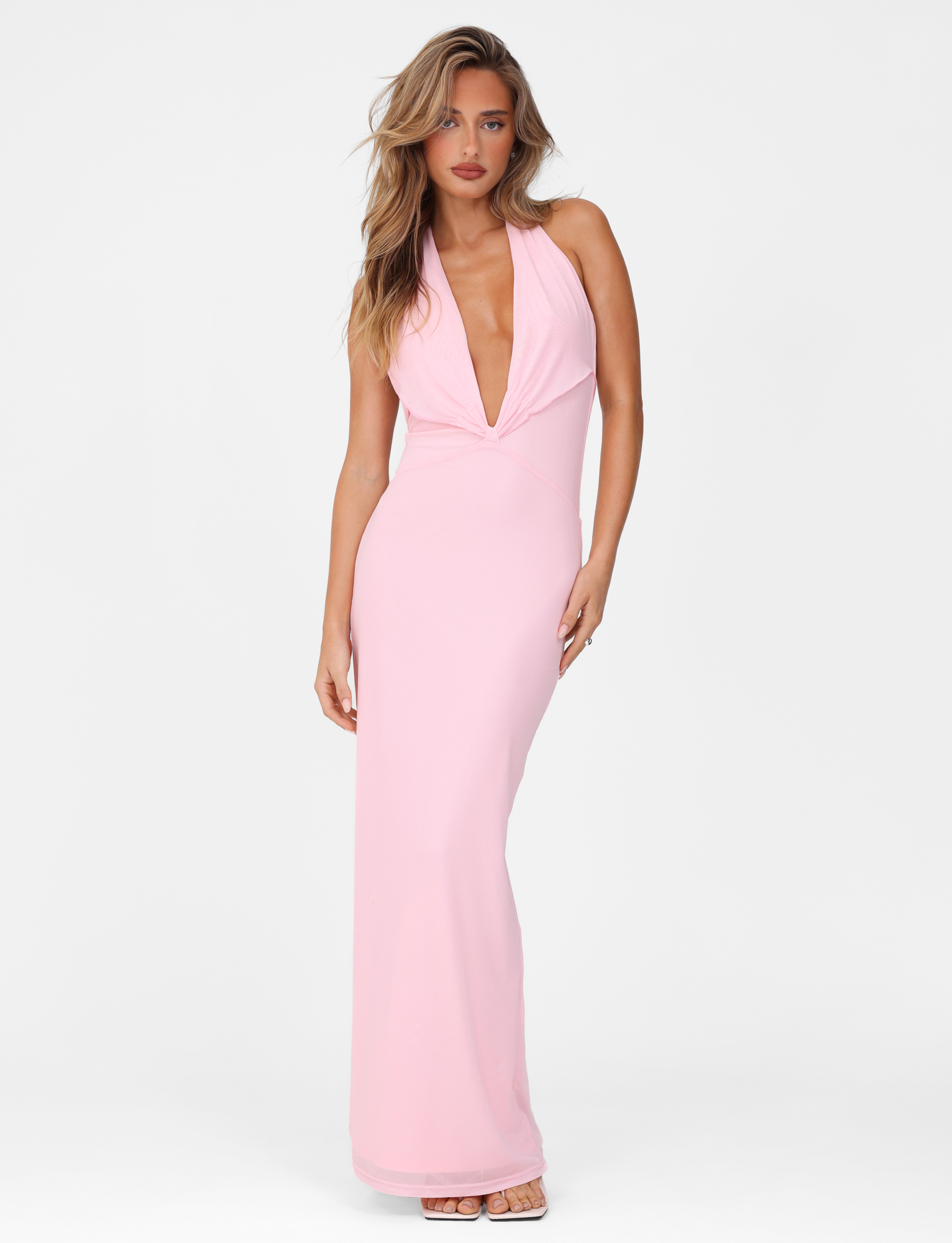 A woman models the TIGERMIST CRESSIDA MAXI DRESS in baby pink, featuring a plunging neckline, standing against a white background with one hand by her side and the other on her hip.