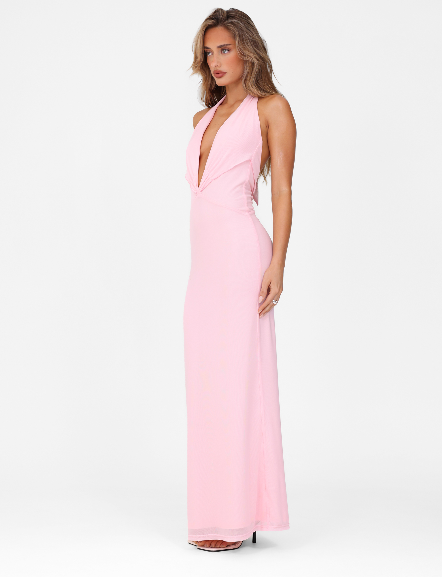 A woman stands against a white background, looking to the side in the TIGERMIST CRESSIDA MAXI DRESS - BABY PINK, a sleeveless halter neck dress with a deep V plunging neckline and maxi length.