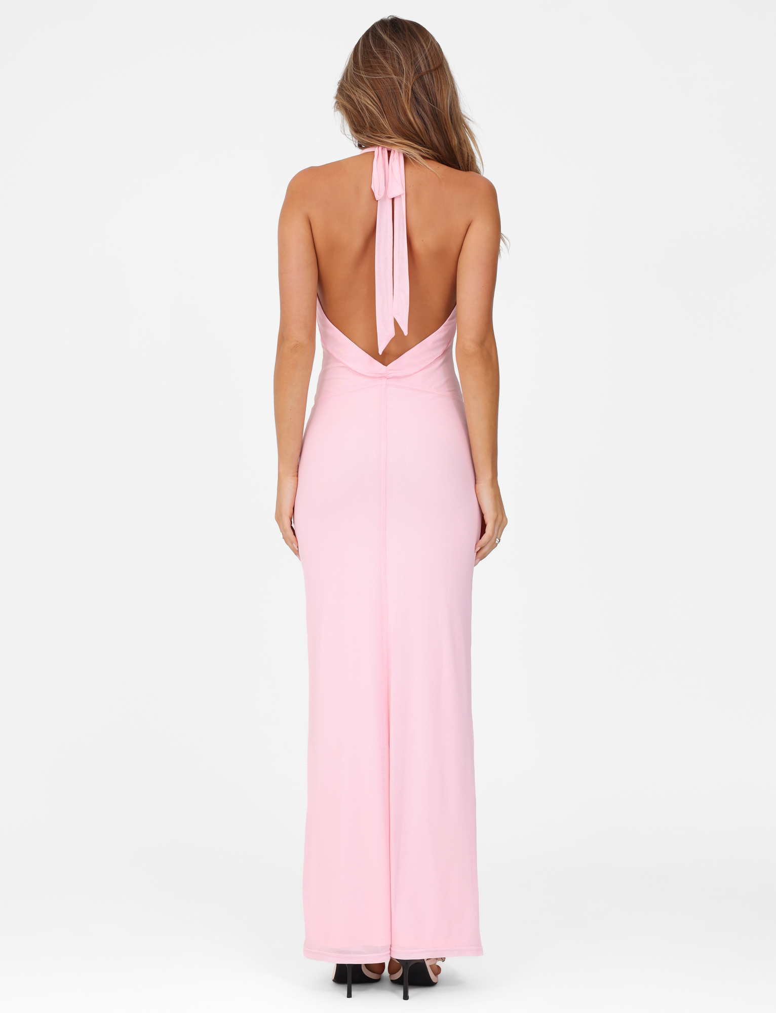 A woman with long, wavy hair models the TIGERMIST CRESSIDA MAXI DRESS in baby pink, standing with her back to the camera in black heels against a plain white background.