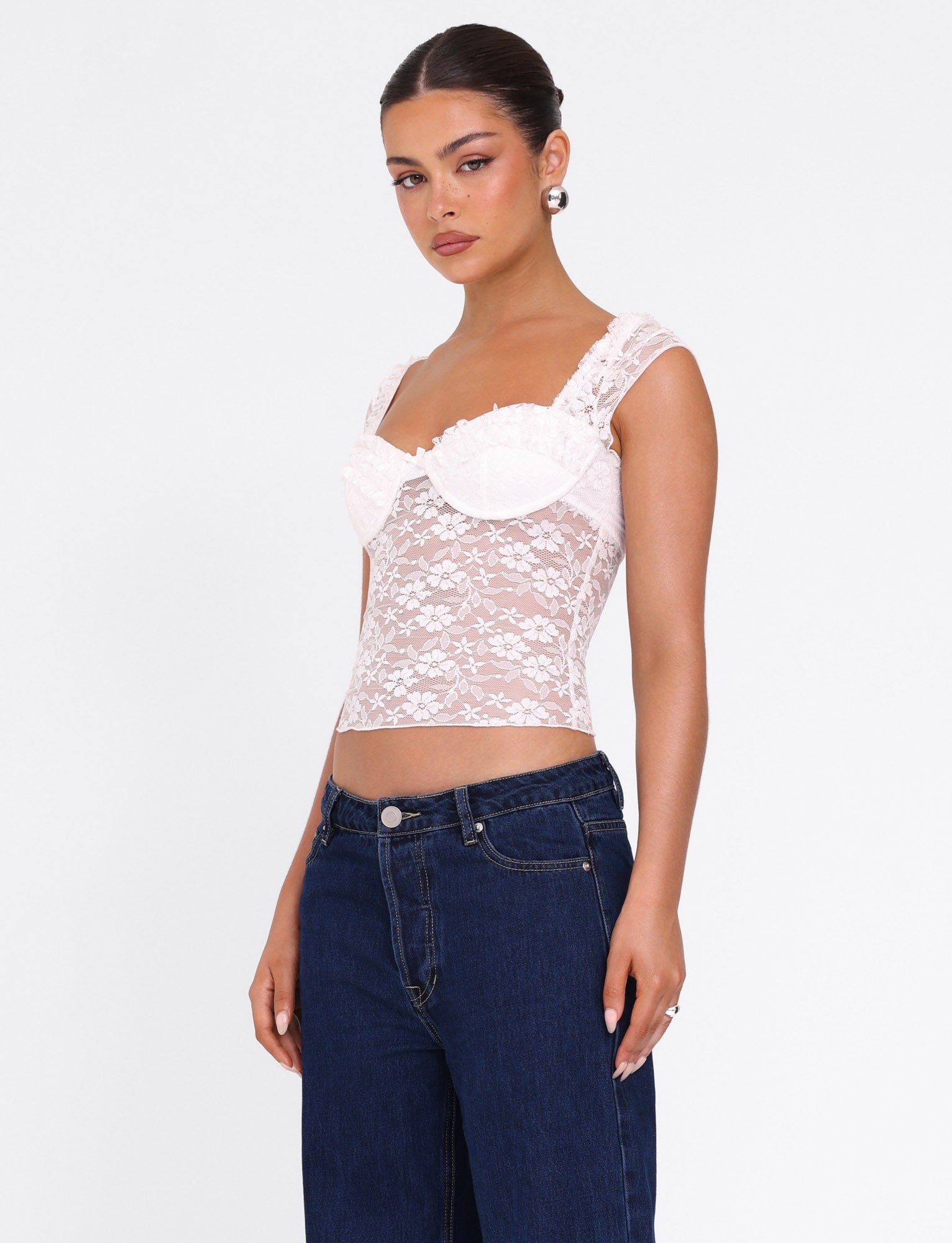 A woman with her hair pulled back and a neutral expression wears the TIGERMIST NYRA TOP in white, a fitted, semi-sheer bustier with lace details, paired with dark blue high-waisted jeans against a plain white background.