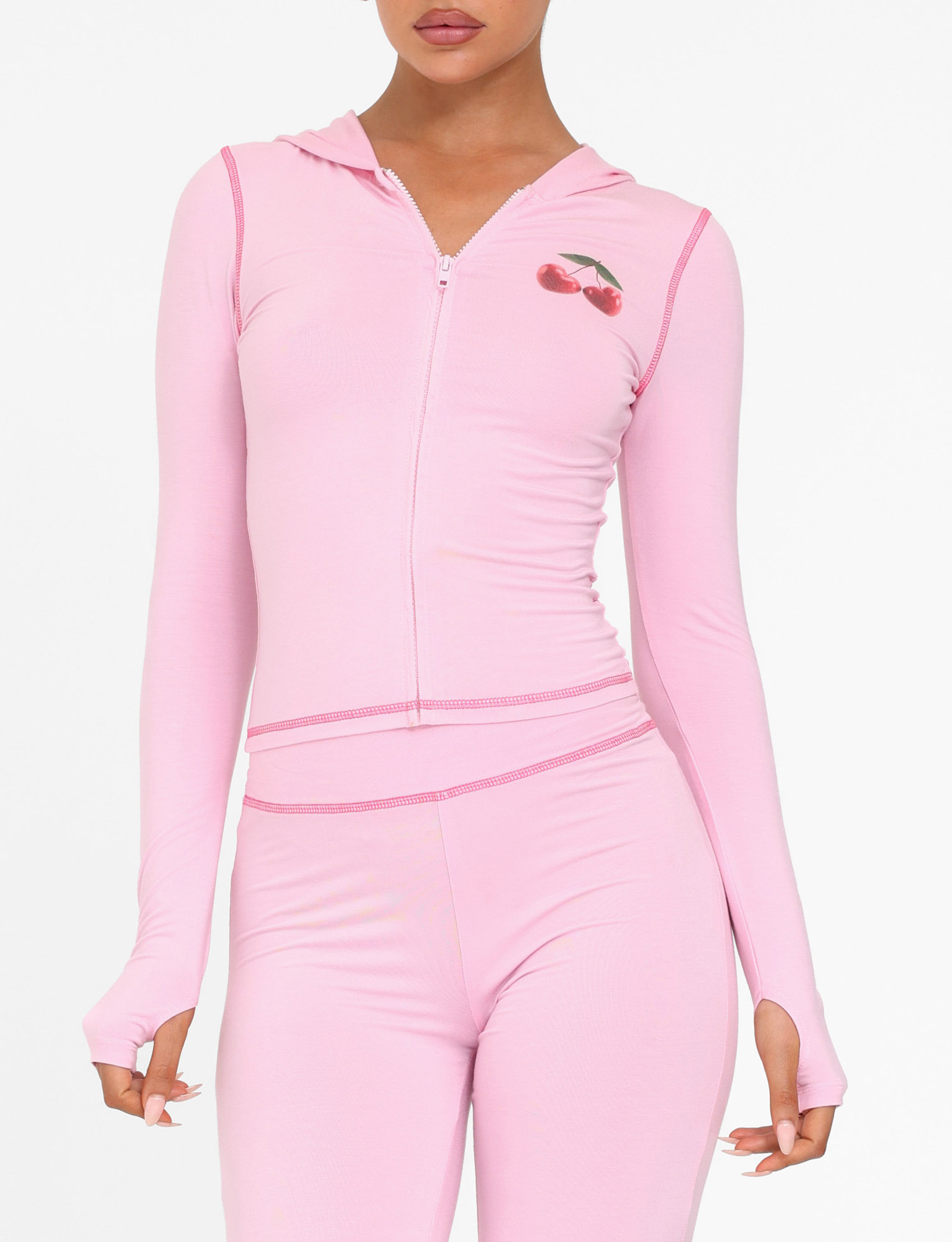 A woman wears the TIGERMIST BLAYZE HOODIE in soft pink with a cherry graphic, hood, thumb holes, and matching high-waisted pants.