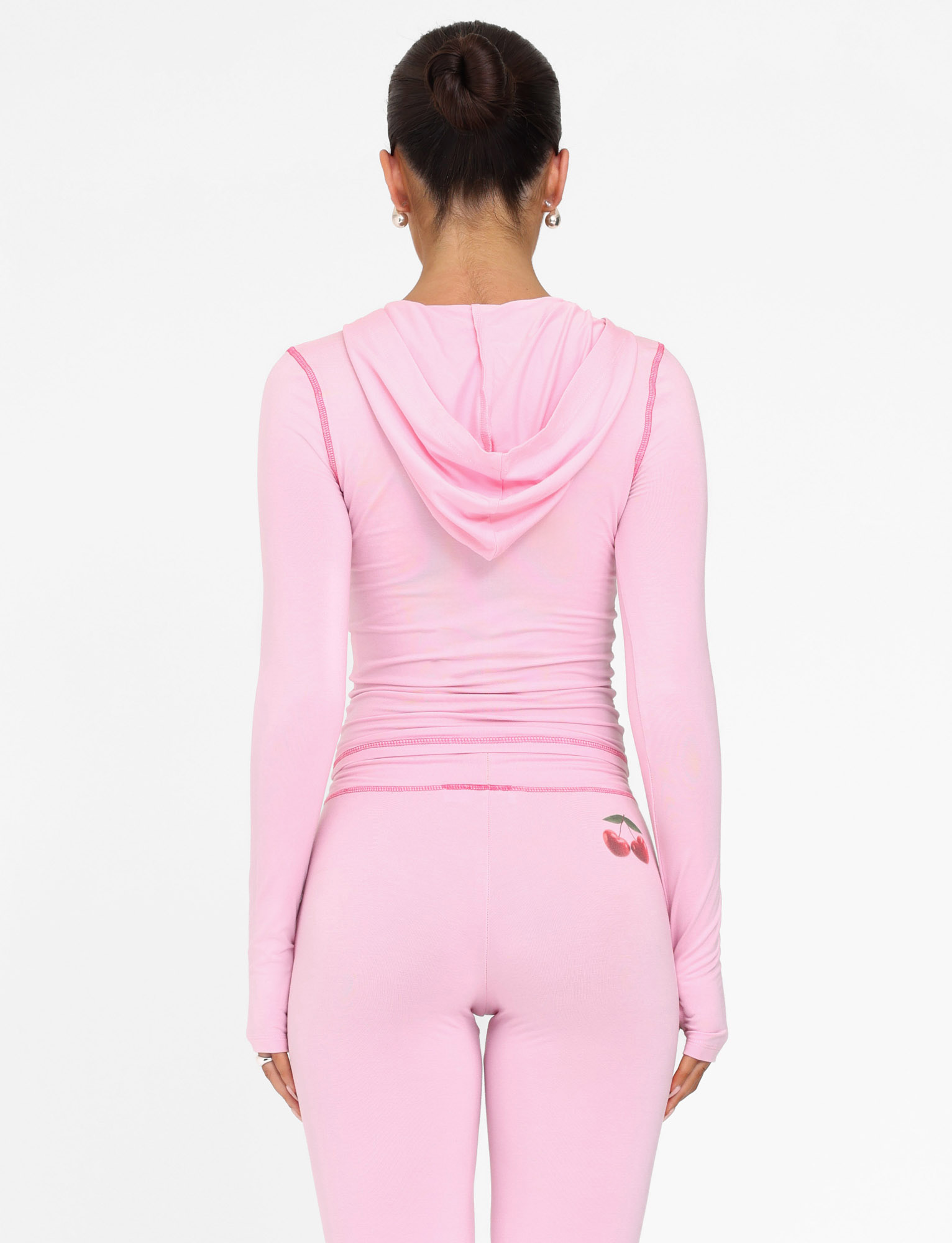 A woman with her dark hair in a bun wears the TIGERMIST BLAYZE HOODIE - PINK: SOFT PINK, paired with matching leggings featuring a small cherry detail at the waistband. She faces away against a plain white background.