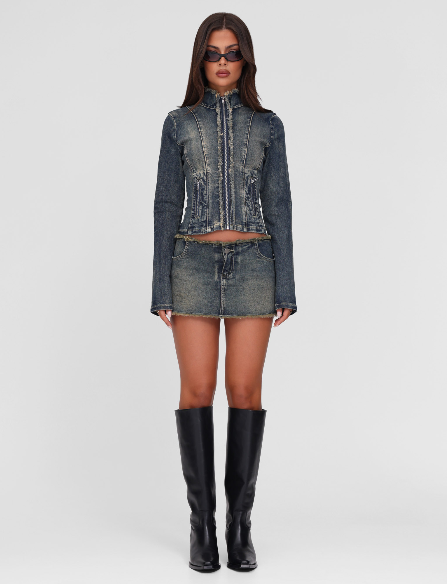 A woman faces forward against a plain background, wearing the TIGERMIST OPHELIE JACKET in denim blue with a matching mini skirt, black knee-high boots, and sunglasses; her arms rest by her sides.