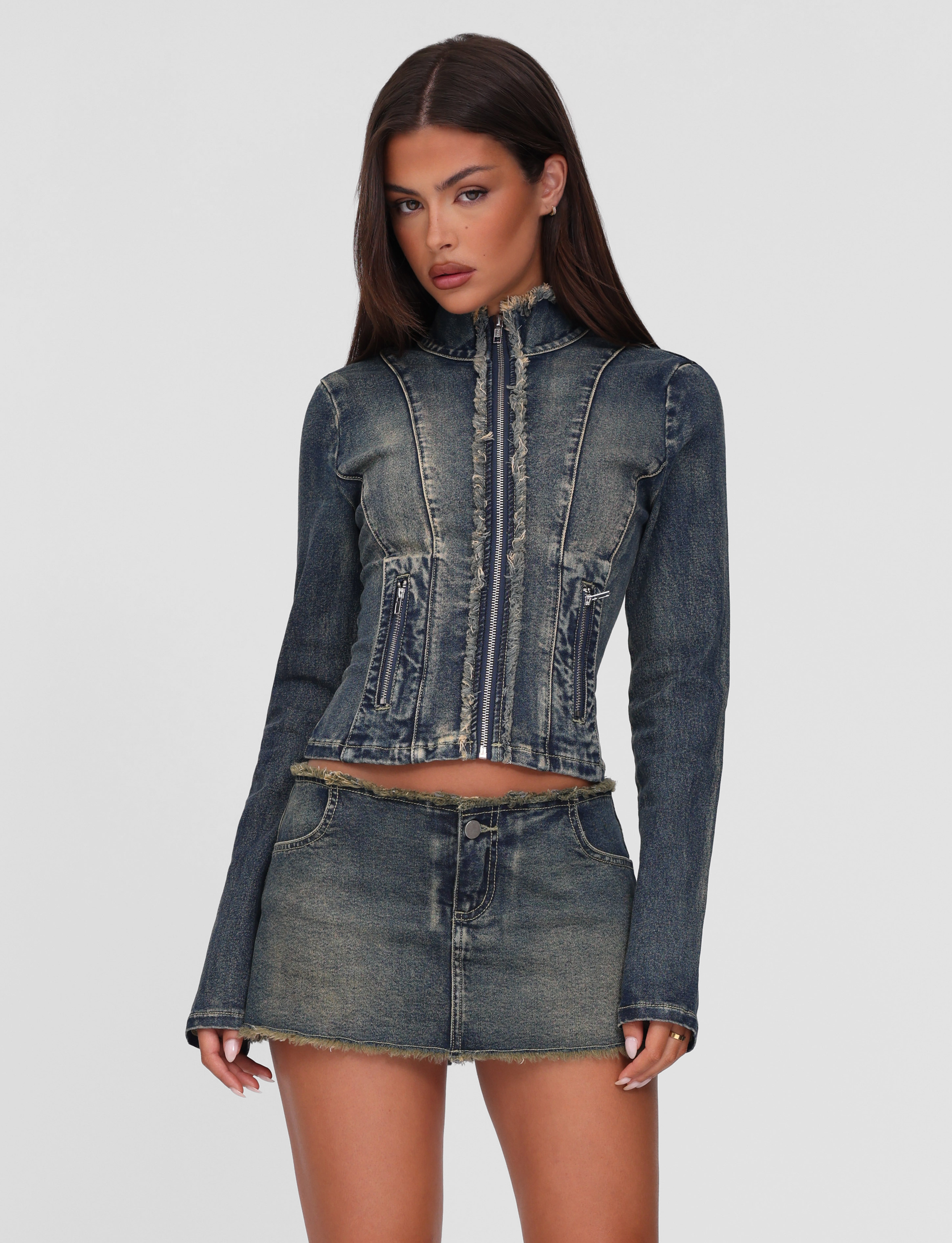 A woman stands against a plain light gray background wearing the TIGERMIST OPHELIE MINI SKORT in denim blue, paired with a relaxed fit, distressed denim jacket featuring a front zipper and frayed details.