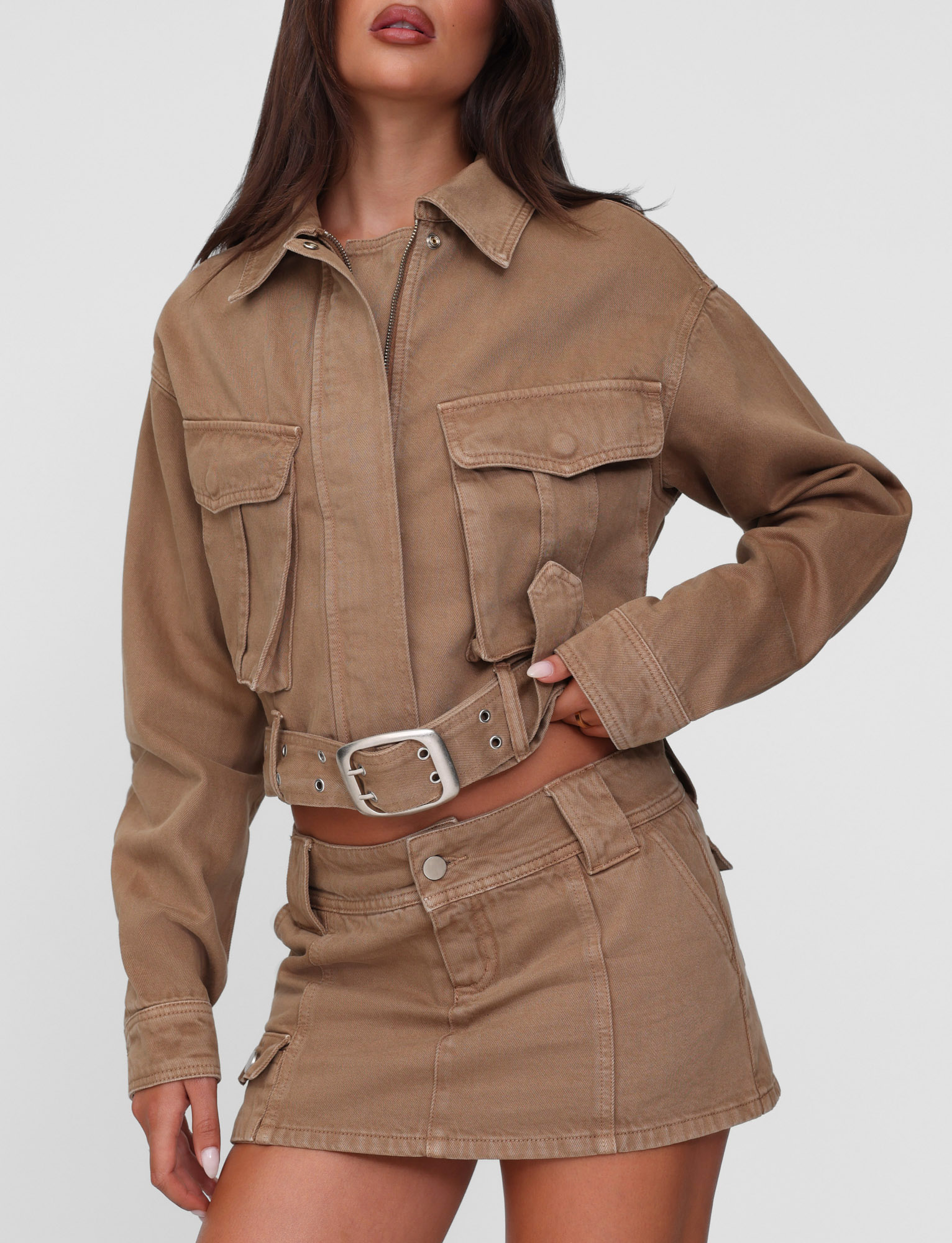 A woman wears the TIGERMIST ZELIE JACKET in washed tan, a neutral cropped utility style with patch pockets, paired with a matching high-waisted mini skirt featuring a silver buckle belt, set against a plain light background.