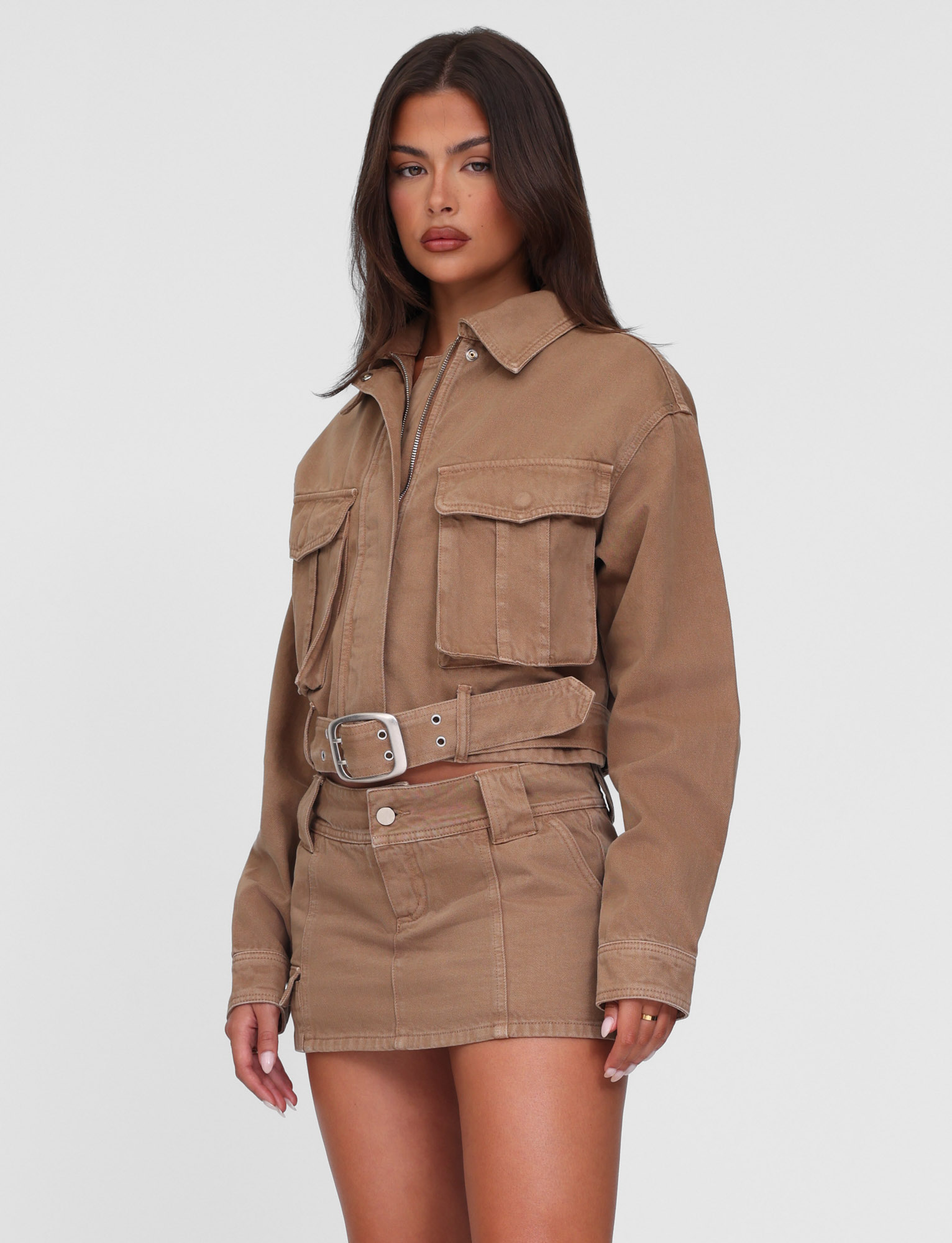 A woman with long dark hair and a neutral expression wears TIGERMIST’s ZELIE MINI SKORT in washed tan sandblast and a matching utility denim jacket, both featuring functional pockets, standing against a white background.