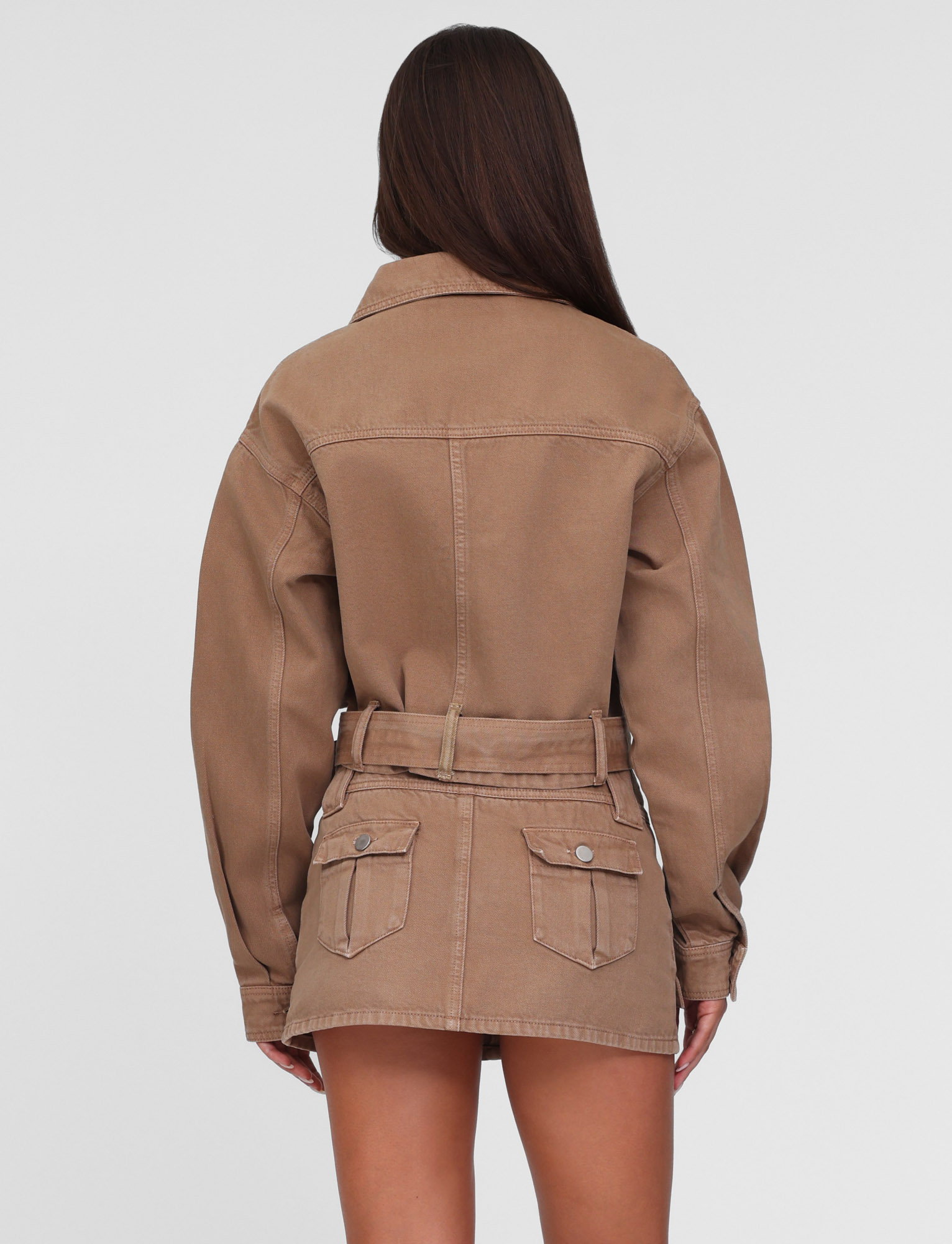 A woman with long brown hair is seen from behind wearing the TIGERMIST ZELIE JACKET in washed tan (neutral sandblast), styled with a matching mini skirt. The utility jacket features patch pockets and a belt. Background is plain white.