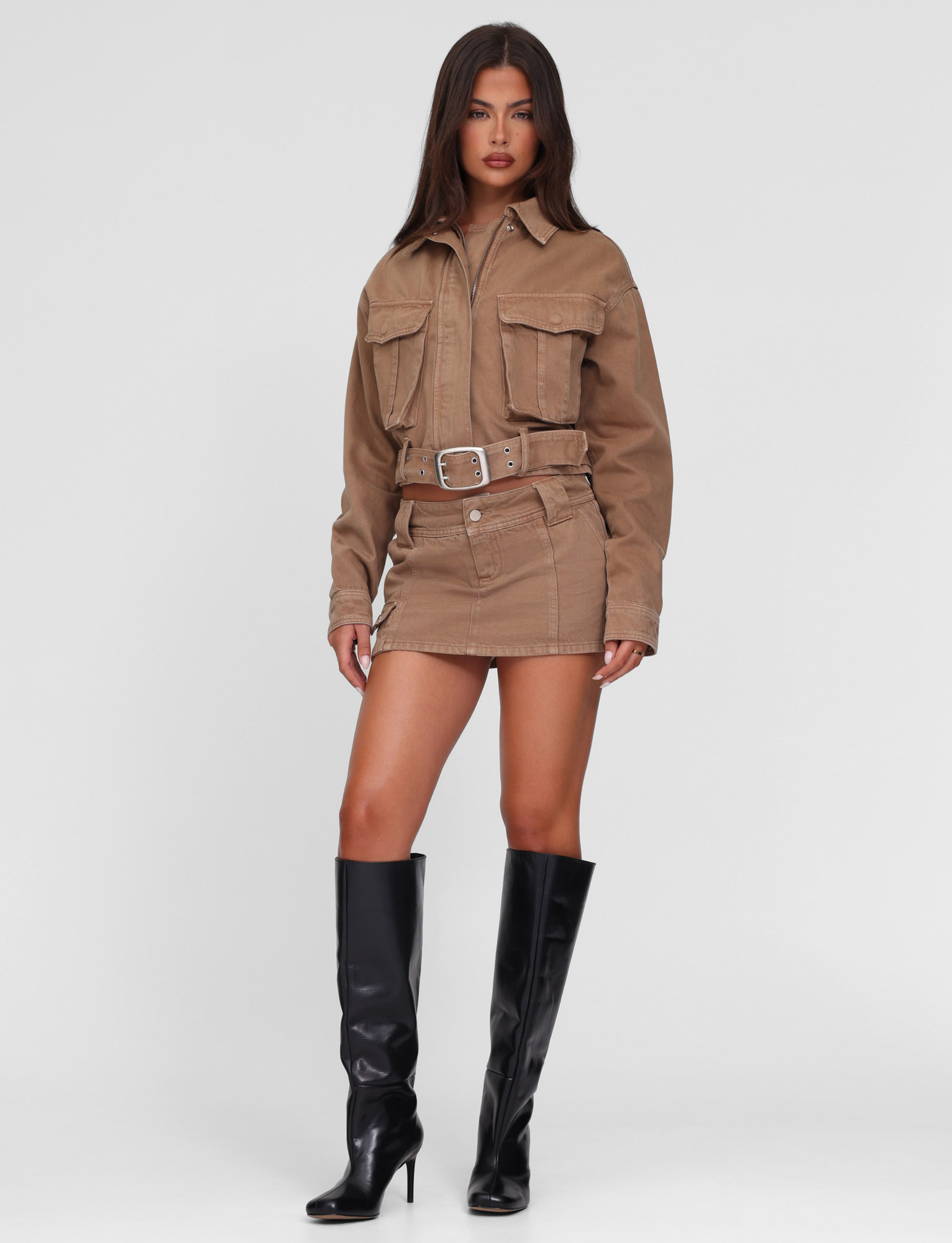 A woman with long straight hair and a neutral expression wears the TIGERMIST ZELIE JACKET - NEUTRAL : SANDBLAST : WASHED TAN, paired with a matching tan mini skirt and knee-high black heeled boots against a white background.