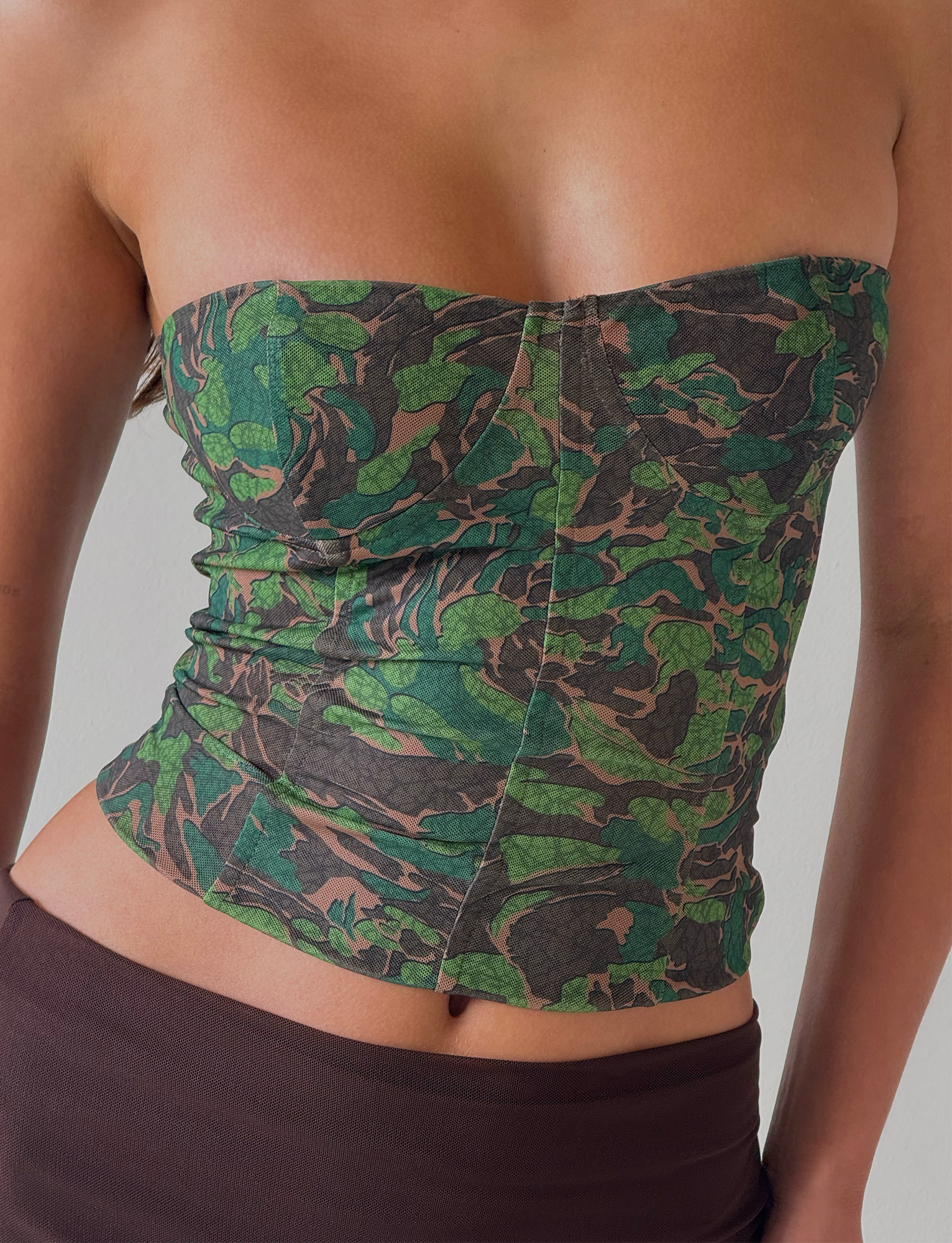 A person wearing the TIGERMIST DULCE CORSET - GREEN : CAMO, a strapless fitted camo top with shaping effect, paired with a brown skirt and standing against a plain light background.