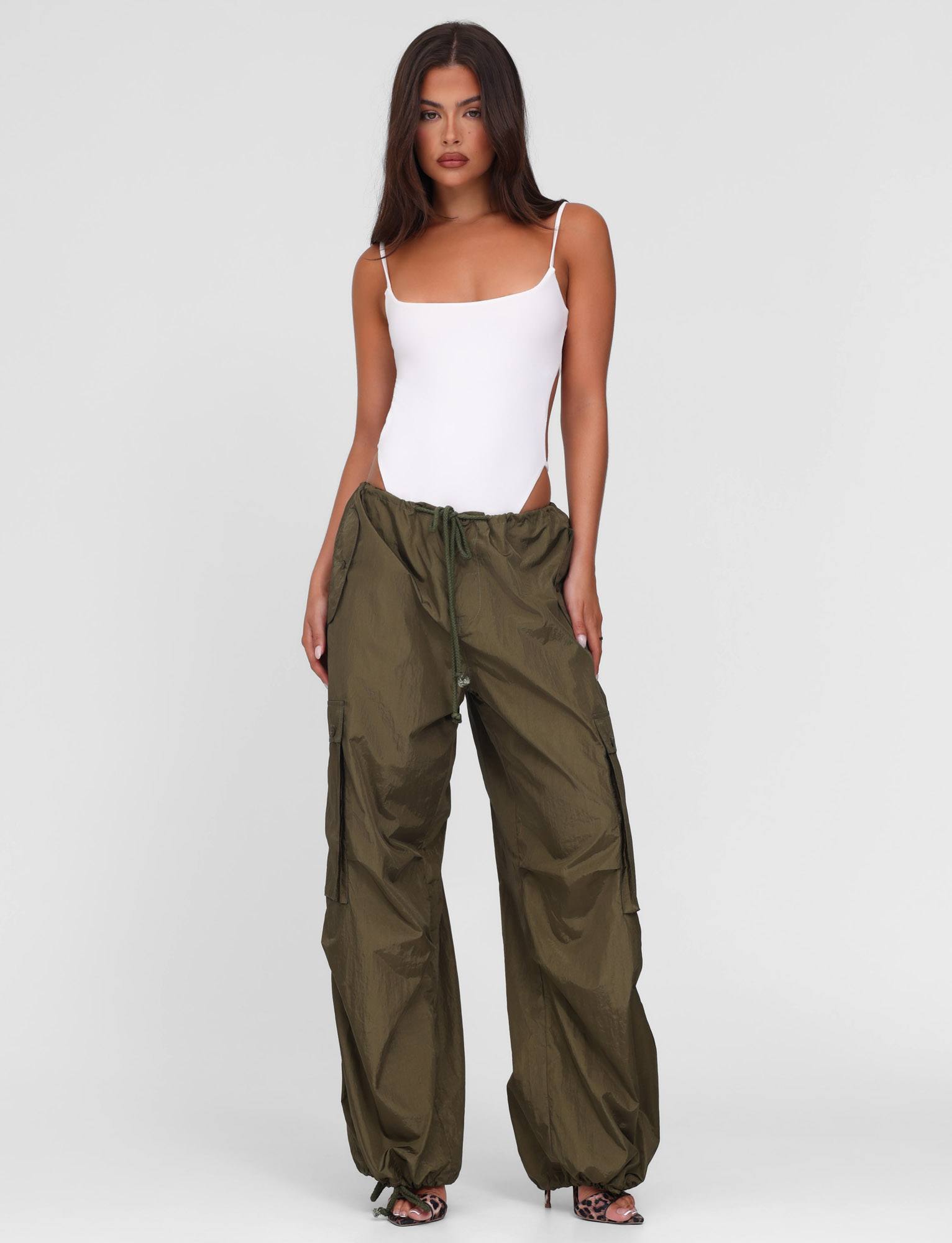 A woman with long dark hair wears a fitted white tank top and TIGERMIST VIOLETTE PANT in khaki green, standing against a plain light gray background.