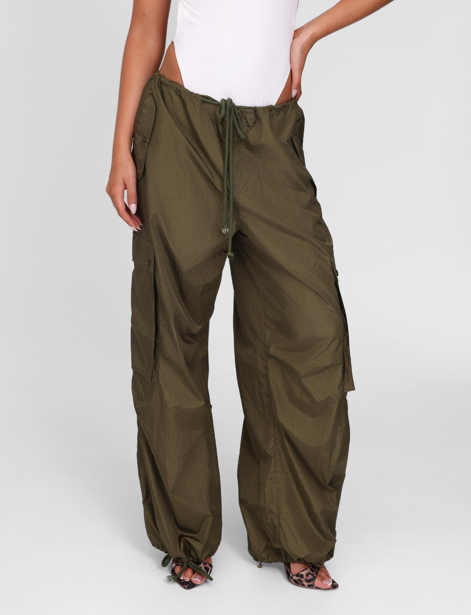 A person wearing TIGERMIST's VIOLETTE PANT in khaki green with adjustable waist and ankle drawstrings, paired with a white sleeveless bodysuit and open-toed heels, stands against a plain gray background.