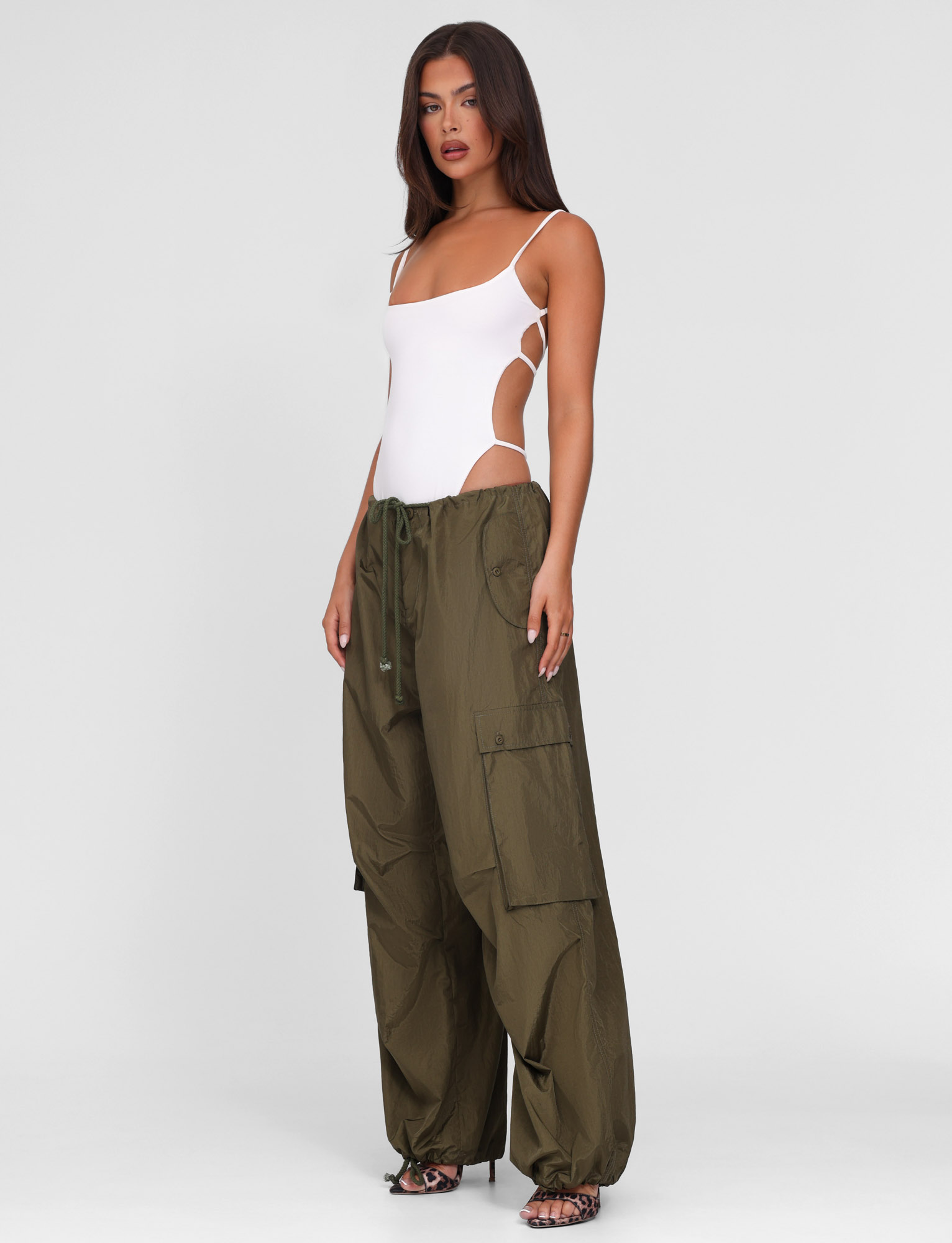 A woman stands against a plain background wearing the TIGERMIST VIOLETTE PANT in khaki green, styled with a white strappy one-piece bodysuit and heeled sandals.