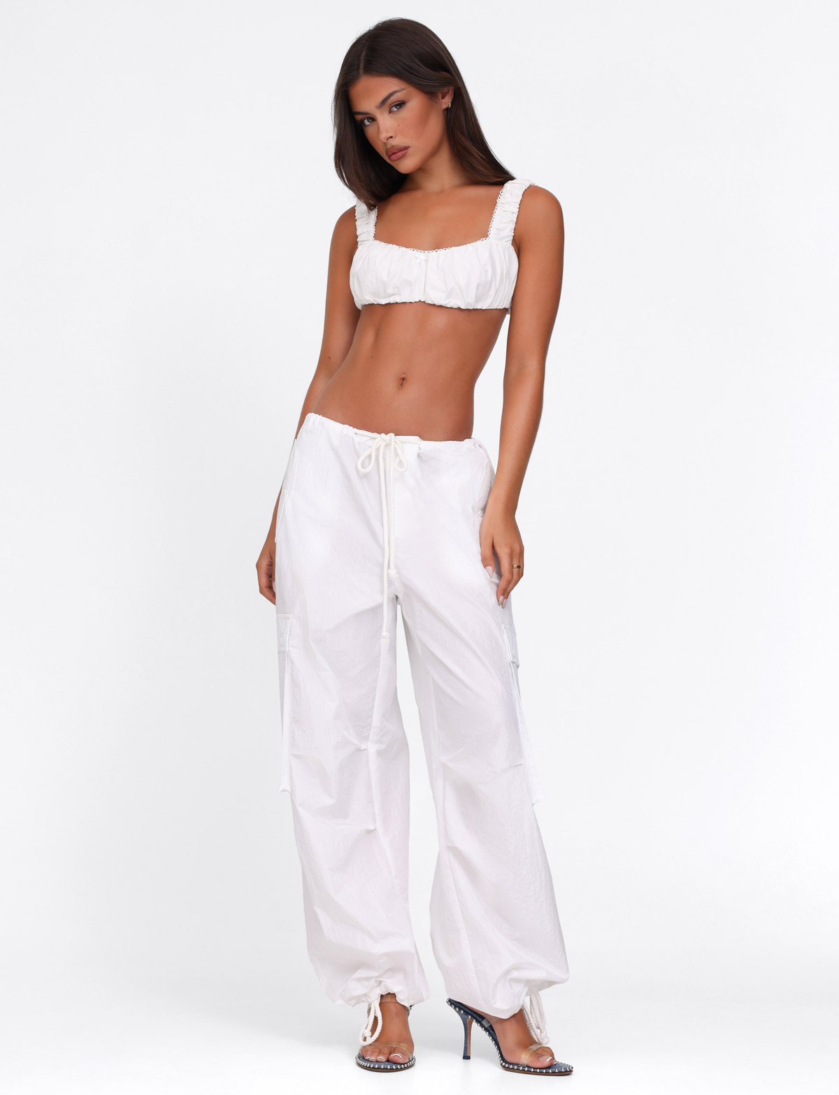Model wears the TIGERMIST VIOLETTE PANT in white—low rise, loose cargo style—paired with a white crop top featuring ruffled straps, standing in high heels against a plain white background.