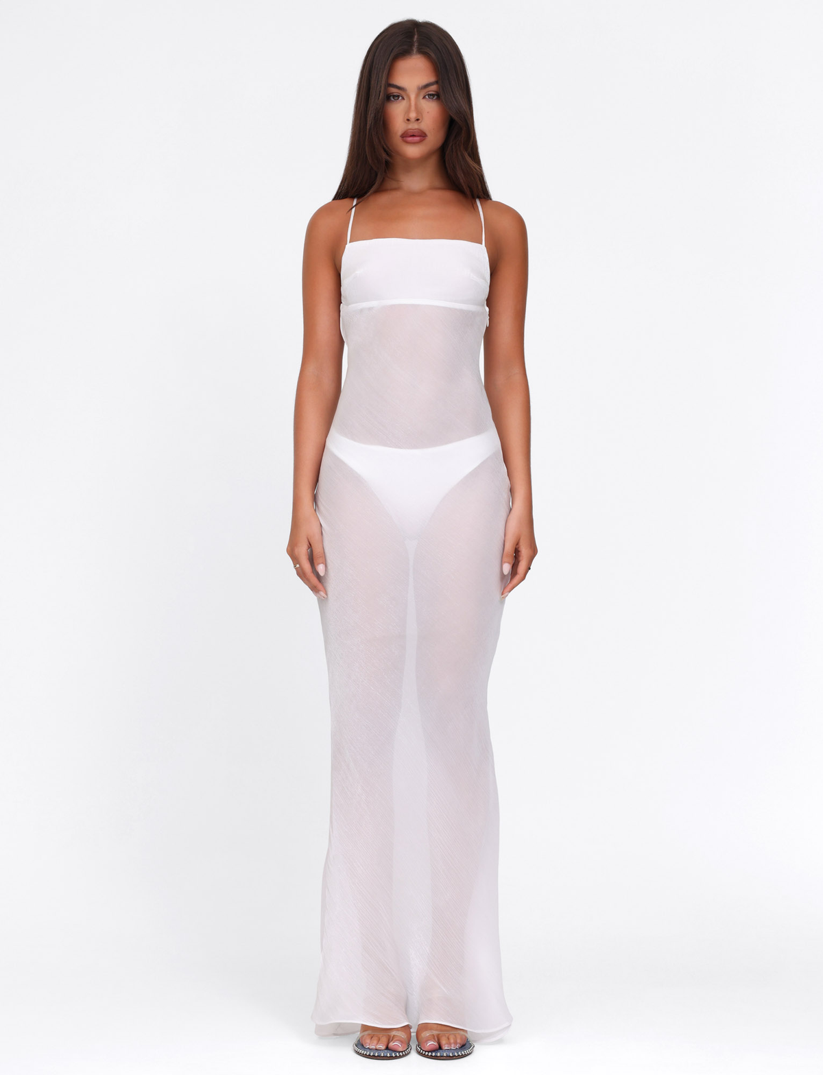 A woman faces forward in the TIGERMIST KIAH MAXI DRESS - WHITE : ICE, a sheer white bias-cut maxi with adjustable crisscross straps, worn over a white bikini-style undergarment and clear high heels, against a plain white background.