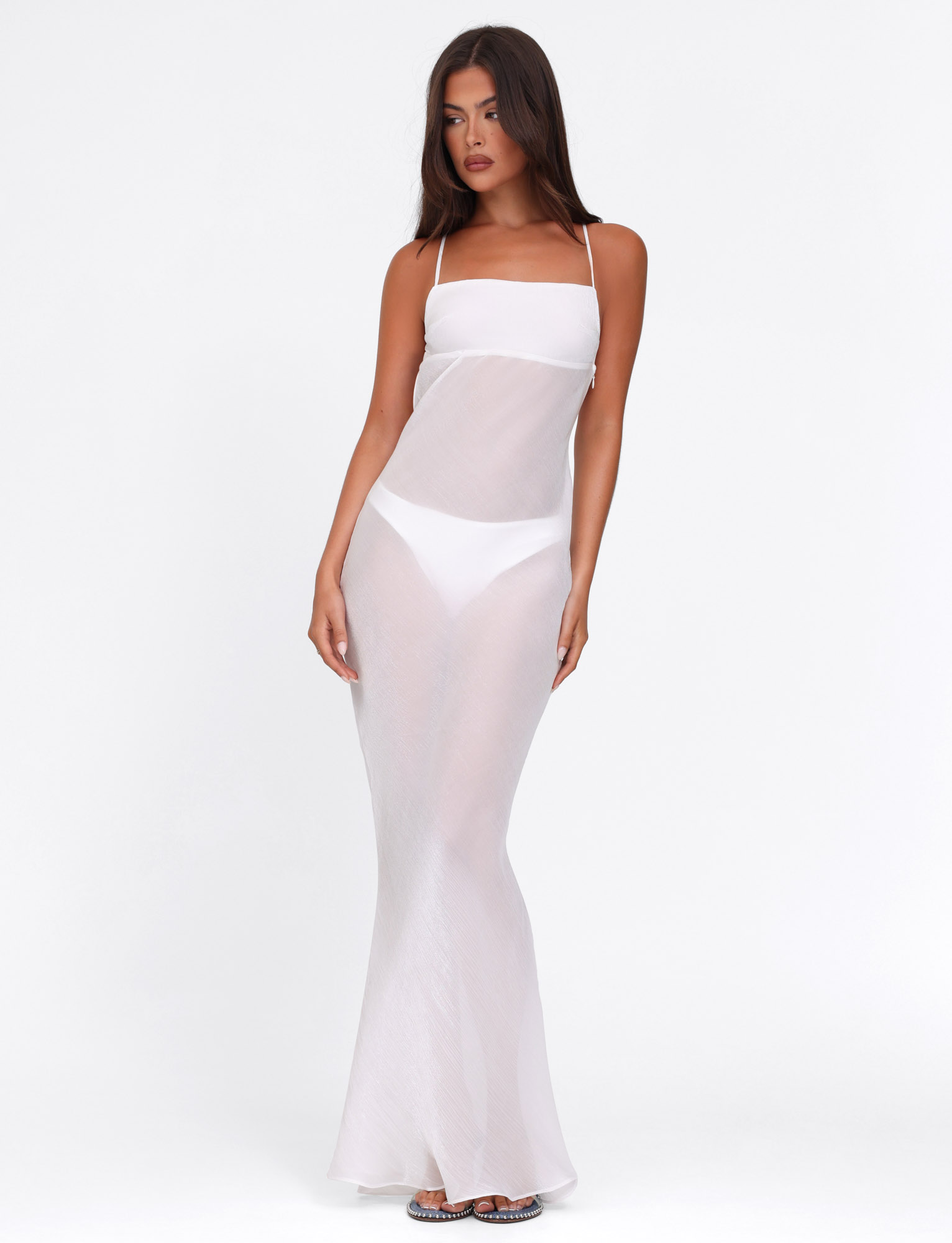 A woman with long, dark hair wears the TIGERMIST KIAH MAXI DRESS - WHITE : ICE, featuring a bias cut and adjustable criss-cross shoulder straps, over a bandeau bra and high-waisted briefs. She stands against a plain white background in open-toed sandals.