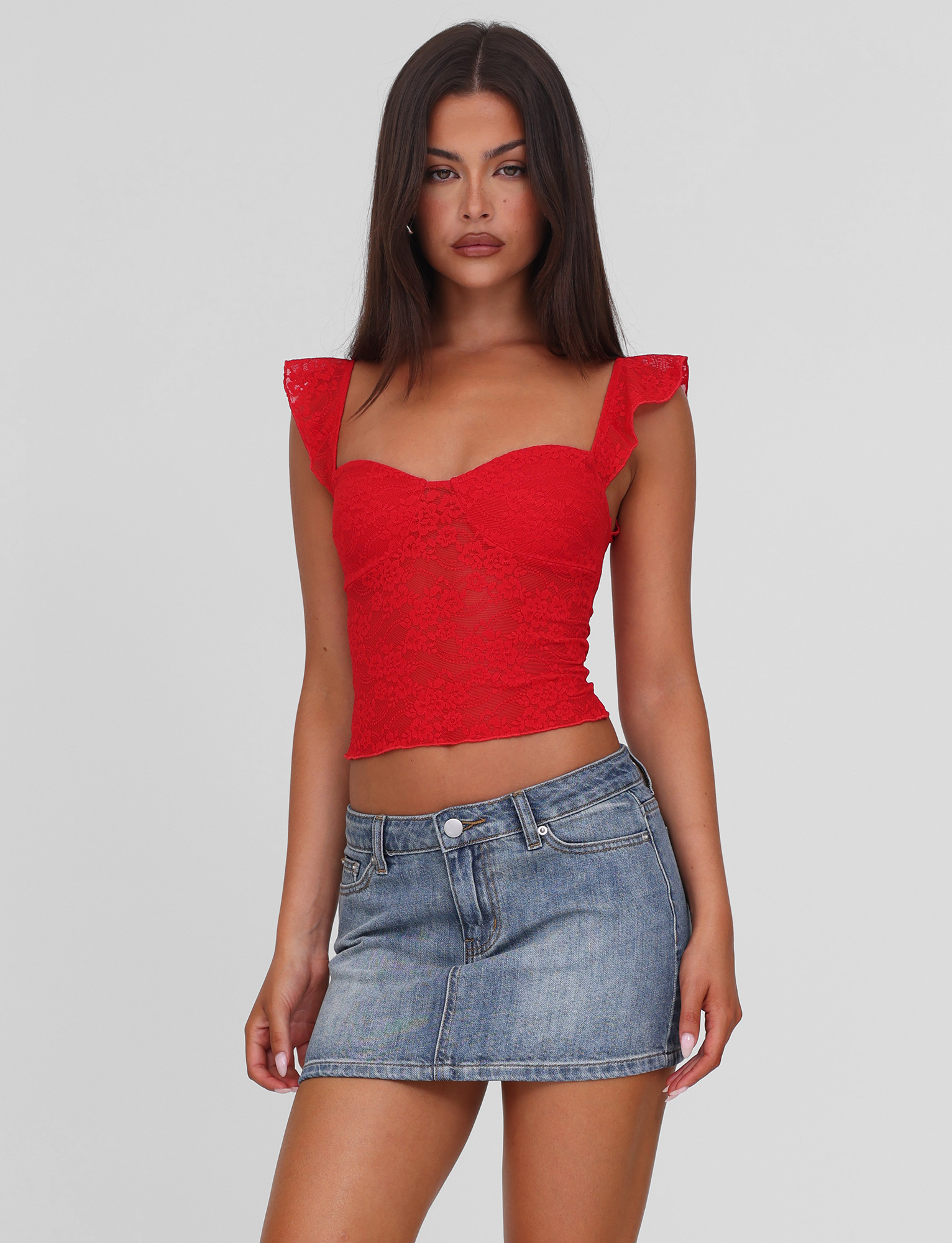 A woman with long dark hair models the TIGERMIST LORNA TOP in red, featuring frilled sleeves, paired with a short blue denim skirt against a plain light gray background.