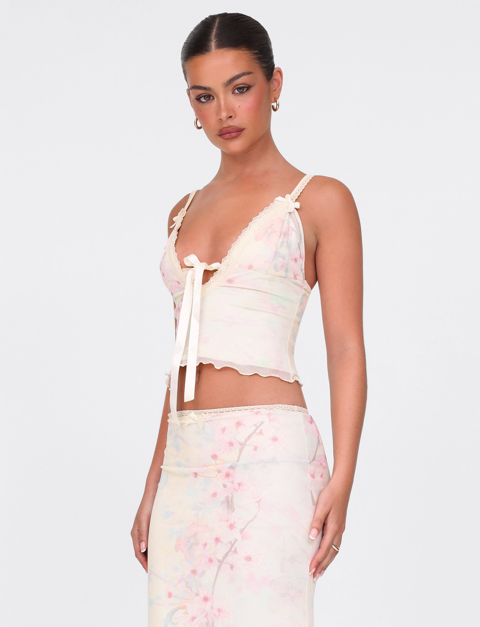 A woman with dark hair in a sleek bun wears the TIGERMIST LONIE TOP in Neutral Floral Washed Cherry Blossom, featuring lace trim and a bow, paired with a matching high-waisted skirt against a plain white background.