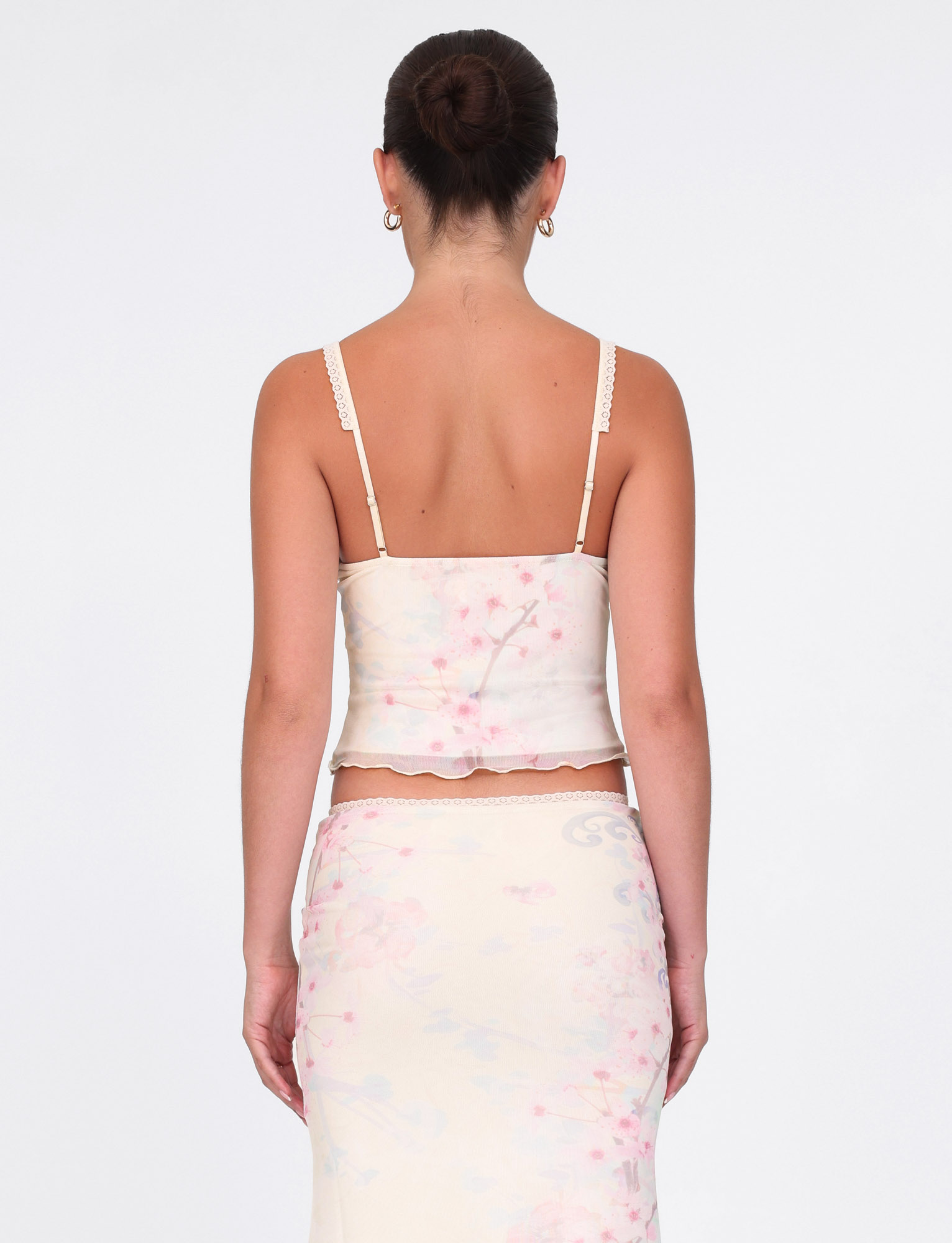 A woman with dark hair in a bun wears TIGERMIST's LONIE TOP in washed cherry blossom floral, featuring lace-trim straps and paired with a matching high-waisted skirt. She accessorizes with gold hoop earrings against a plain white background.