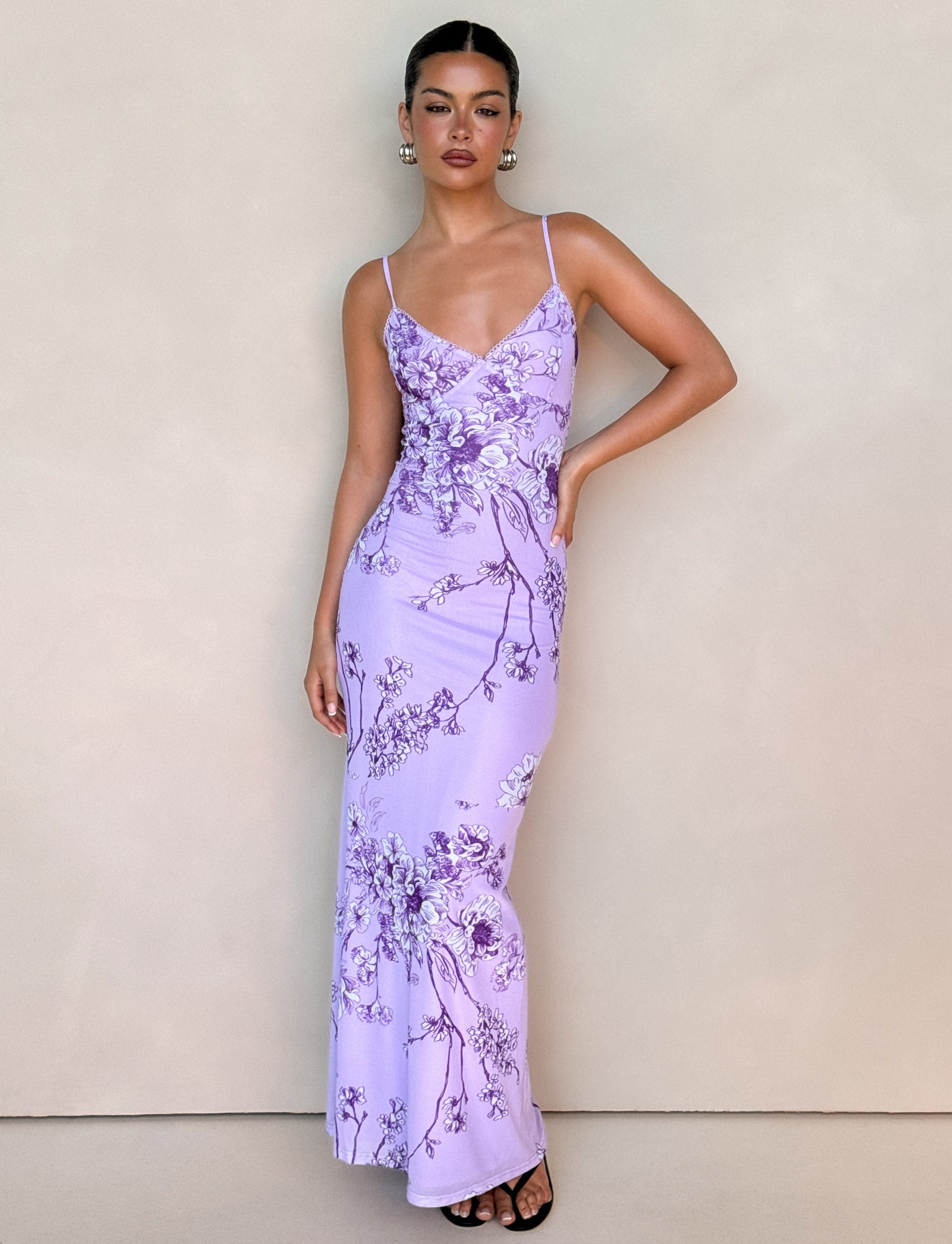 Against a plain backdrop, a person stands confidently wearing the TIGERMIST JULIANNA DRESS in purple ceramic floral with spaghetti straps. Their neat hair and hoop earrings complement the look, paired with black sandals as they pose with one hand on their hip.