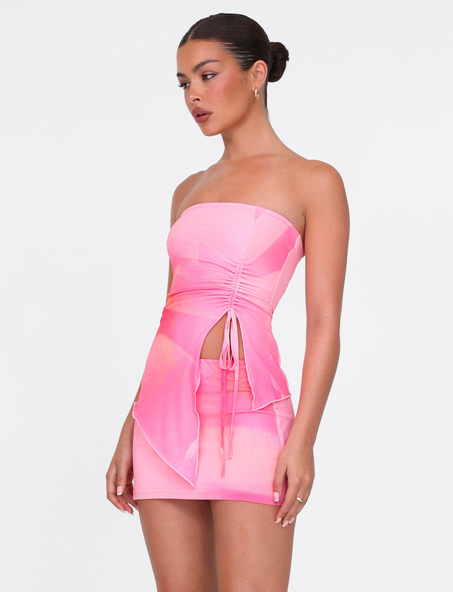 A woman models the TIGERMIST SARIAH TOP in Pink Blossom Macro Floral, featuring a strapless design, ruched front cutout, center tie, and an asymmetrical layered skirt, shown against a plain white background.