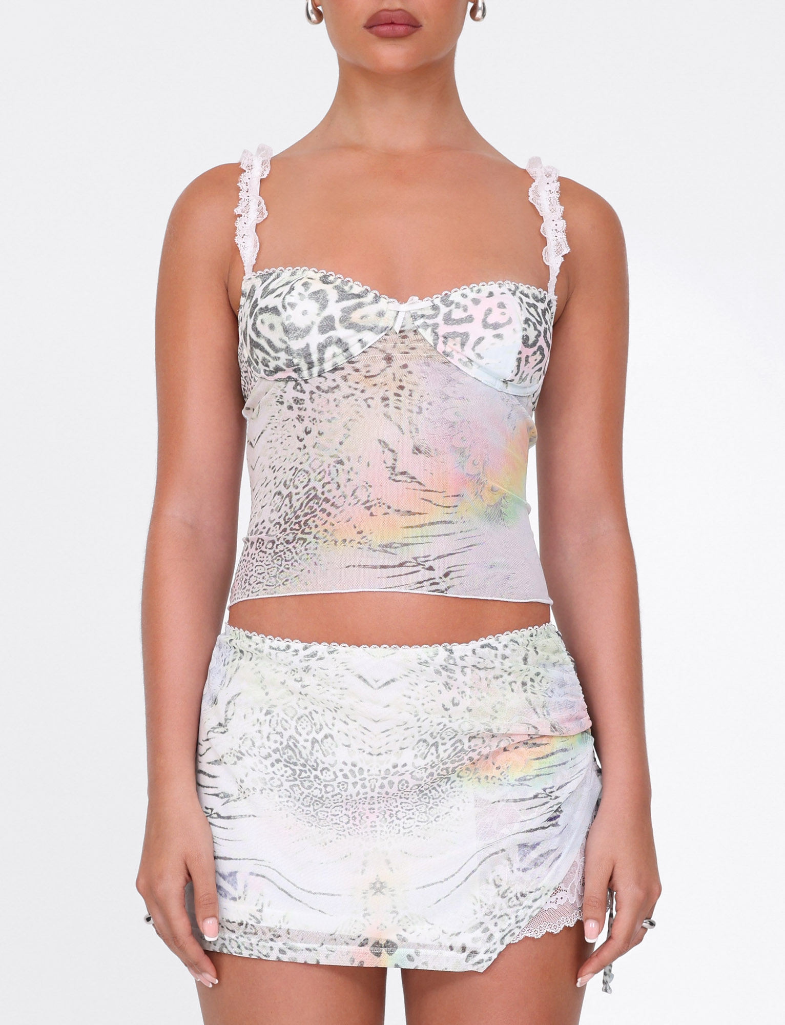 A woman wears the TIGERMIST JOSEPHINE MINI SKIRT in white animal print—featuring ruched side seams and iridescent pastel tones—with a lace-trimmed cami, standing against a plain white background.