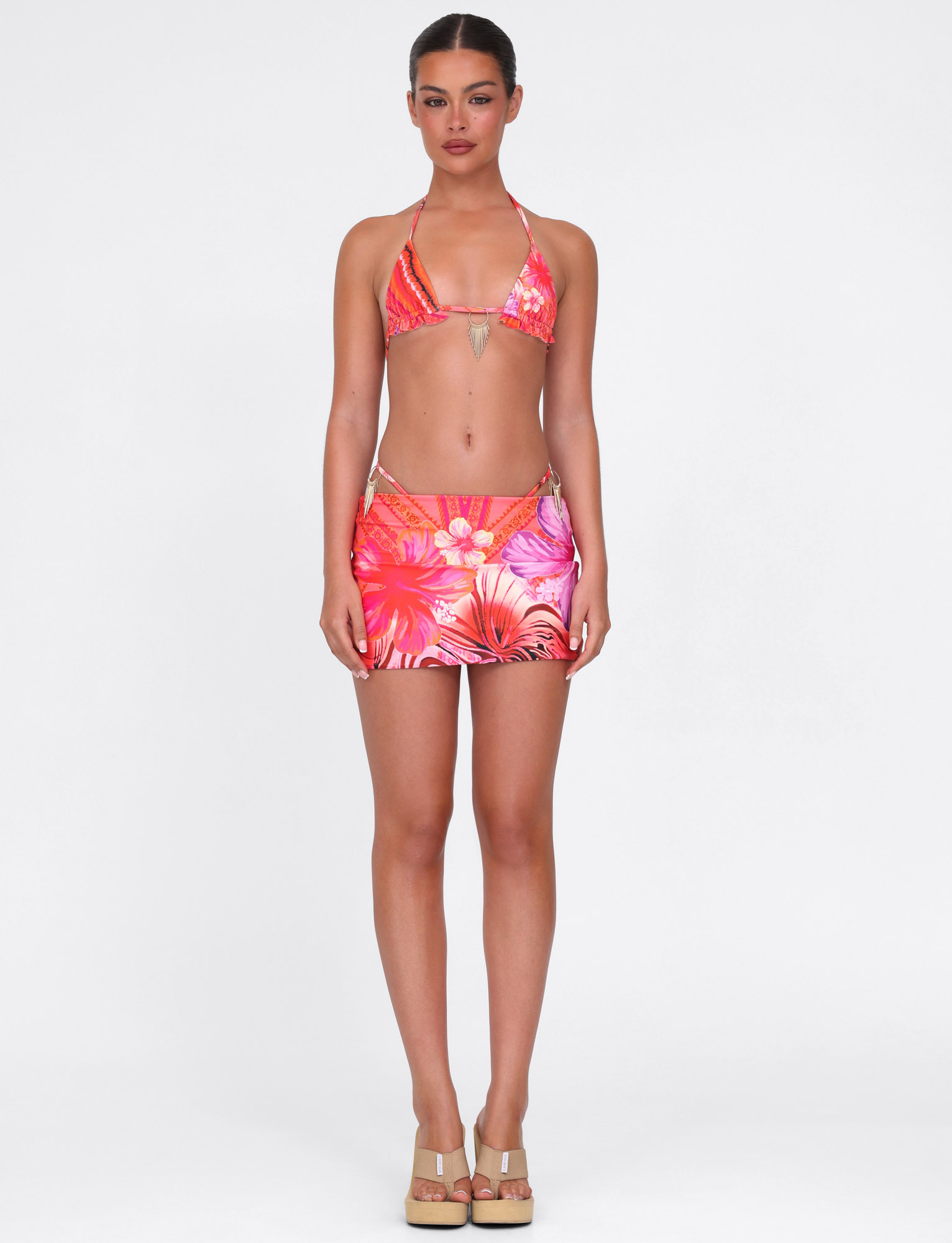 A woman poses against a plain background wearing the TIGERMIST CIANA BIKINI 2PC BOTTOM in Pink Tropical Floral, featuring a matching bikini top and skirt, styled with beige platform sandals.