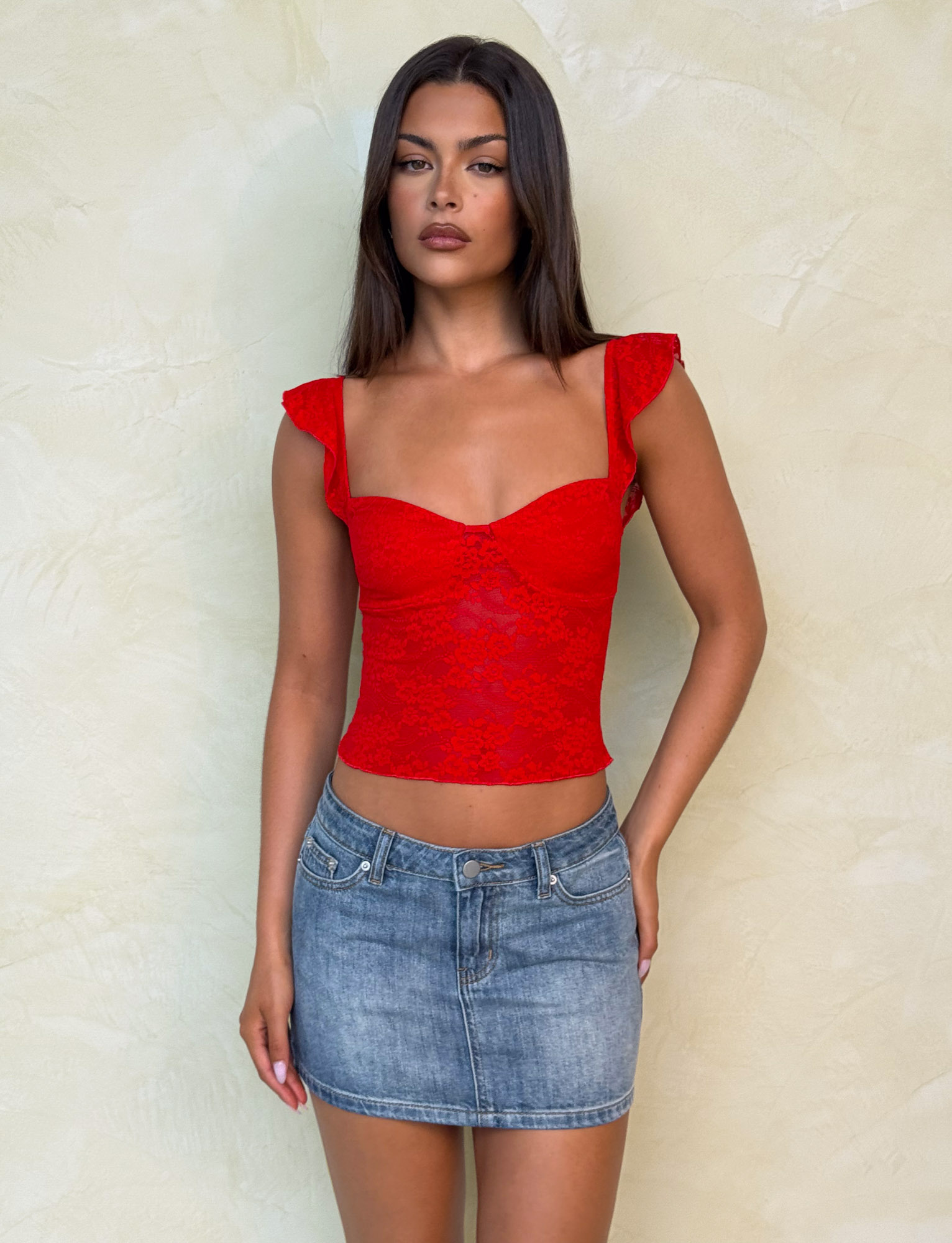 A woman with long, straight dark hair and a neutral expression stands against a light textured wall, wearing a red lace sleeveless top with ruffled straps and the TIGERMIST MICHAYLA MINI SKIRT - MID WASH.