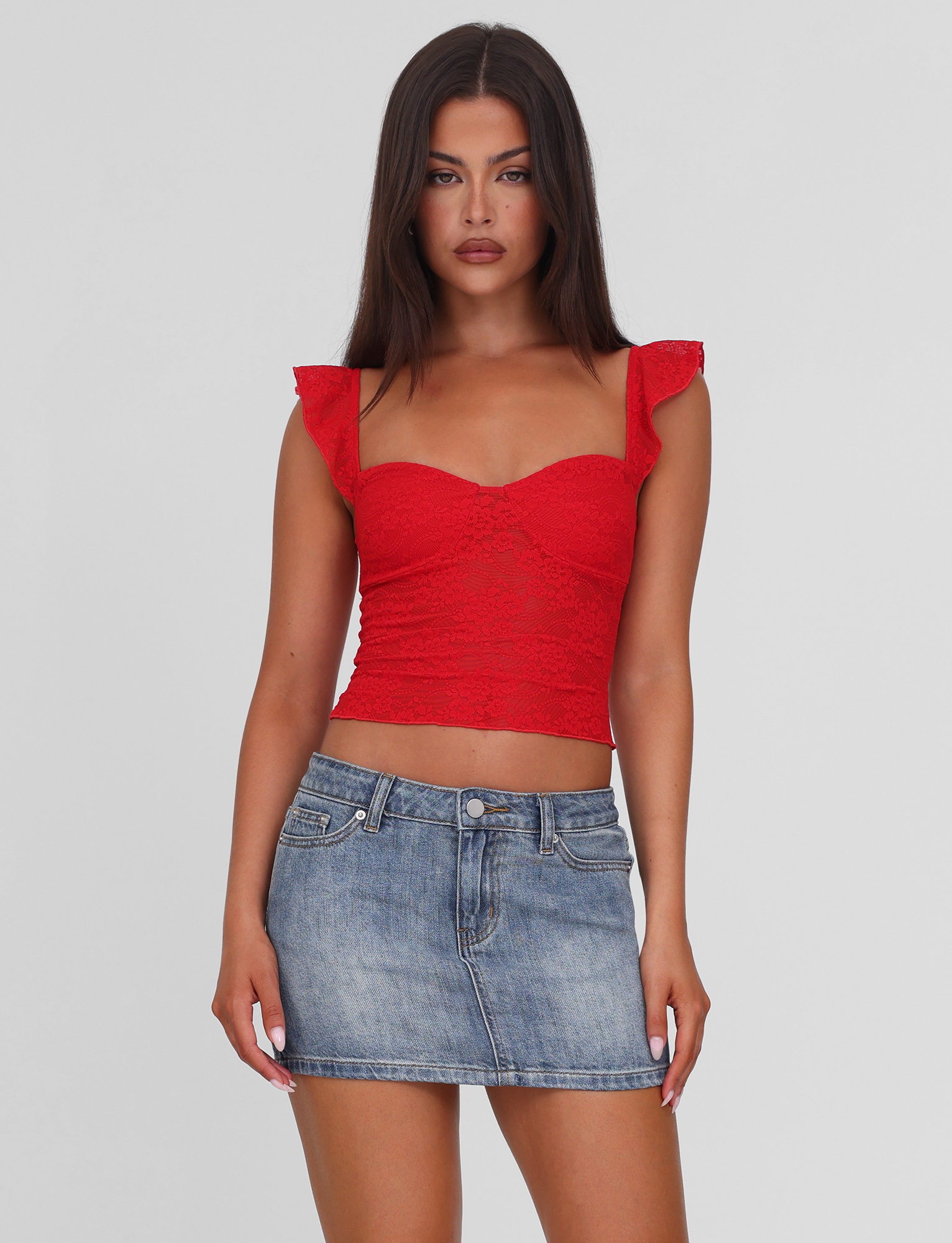A woman with long dark hair models the TIGERMIST MICHAYLA MINI SKIRT - MID WASH, styled with a red lace crop top with ruffled sleeves, against a plain white background.