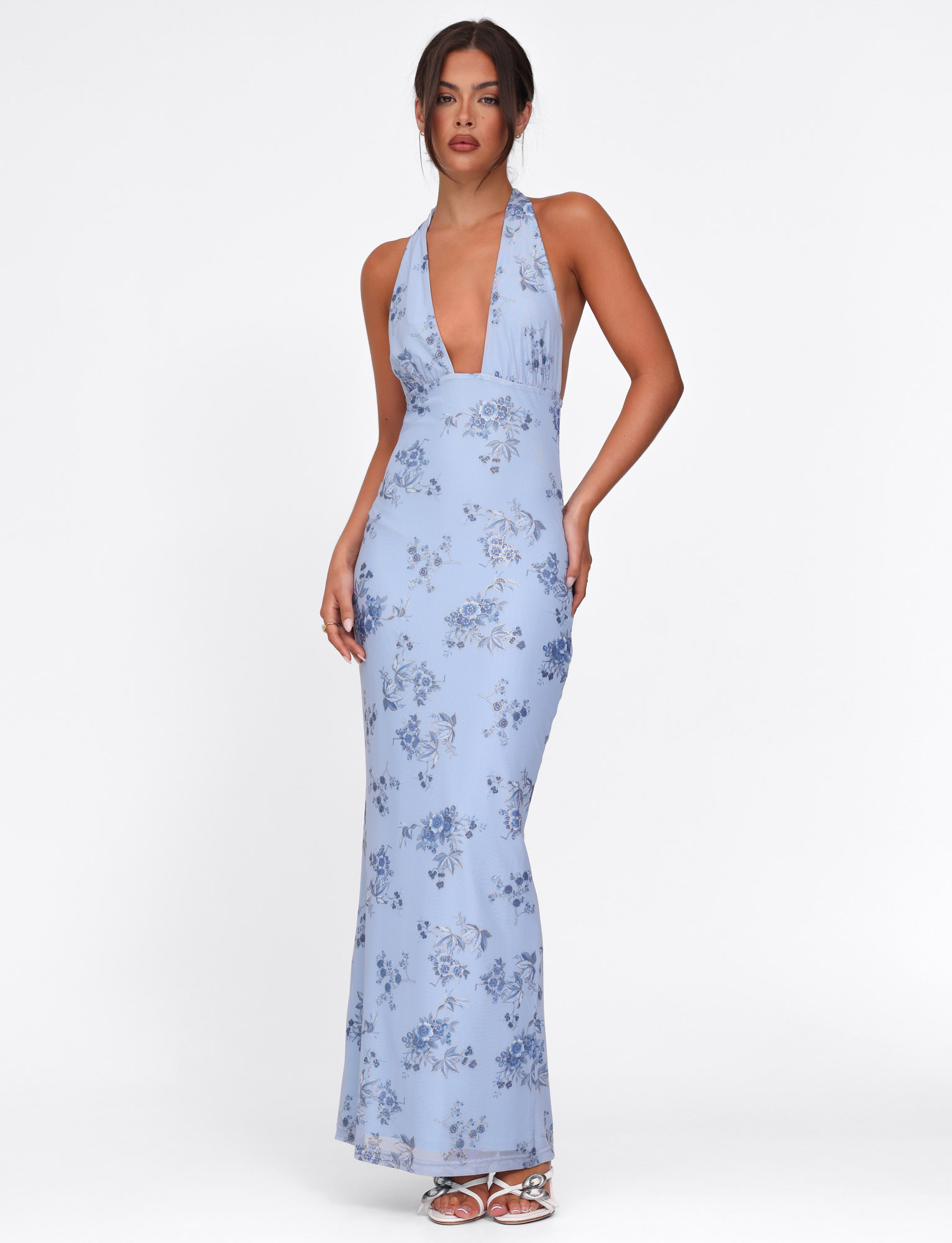 A woman models the TIGERMIST PHAEDRA MAXI DRESS in blue floral, featuring a sleeveless design, deep V-neckline, and metallic foil detail, standing against a plain white background.