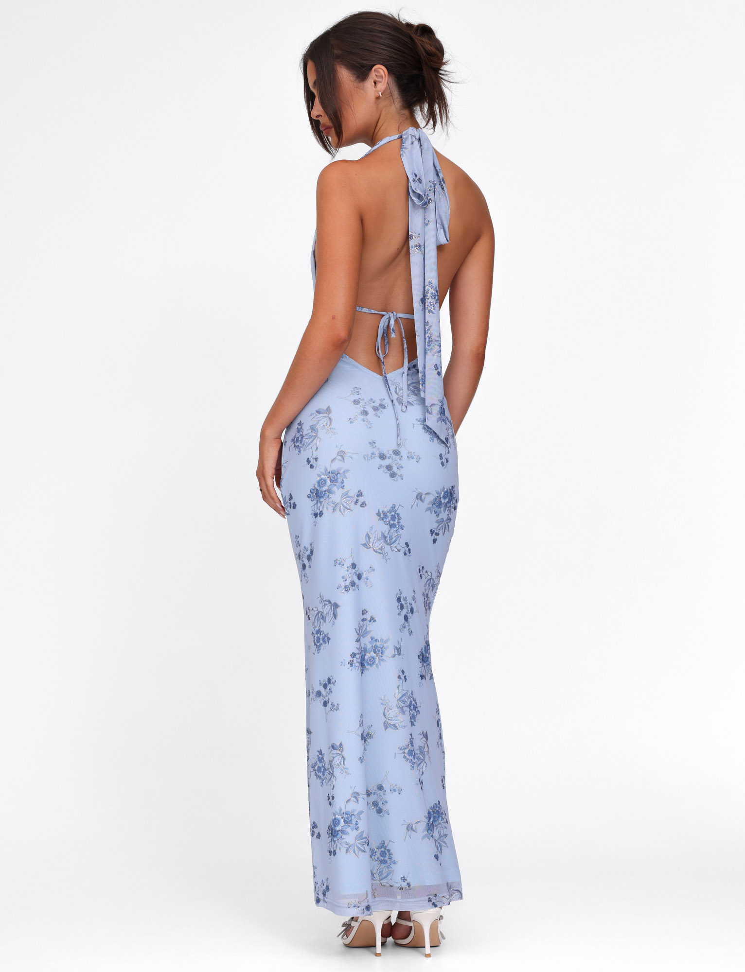 A woman stands sideways in the TIGERMIST PHAEDRA MAXI DRESS - BLUE FLORAL, featuring a backless halter neck with neck tie, styled with white heeled sandals and a loose updo, set against a white background.