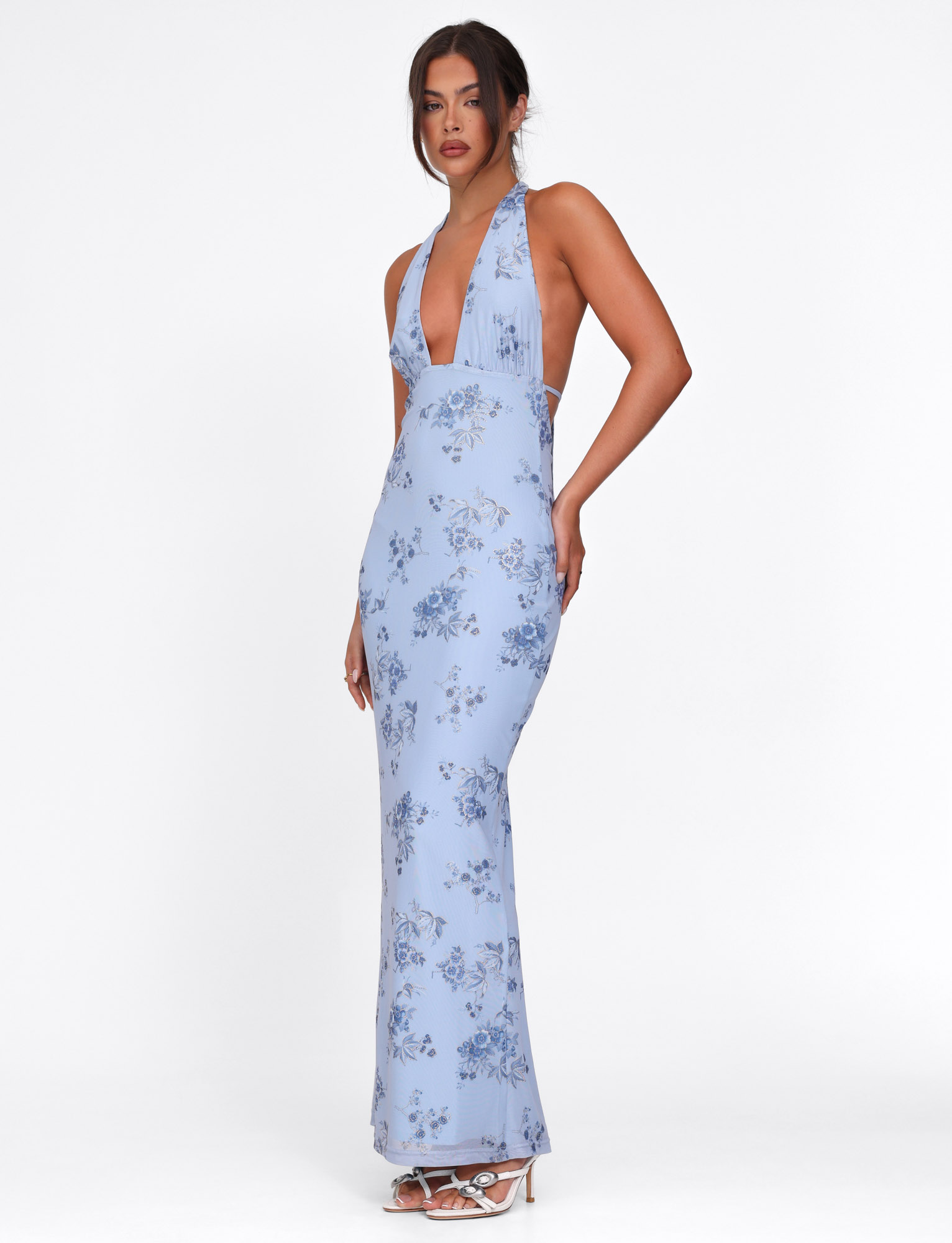 A woman models the TIGERMIST PHAEDRA MAXI DRESS in blue floral with a deep V-neck and halter straps, featuring subtle metallic foil details. She completes the look with white high-heeled sandals against a plain white background.