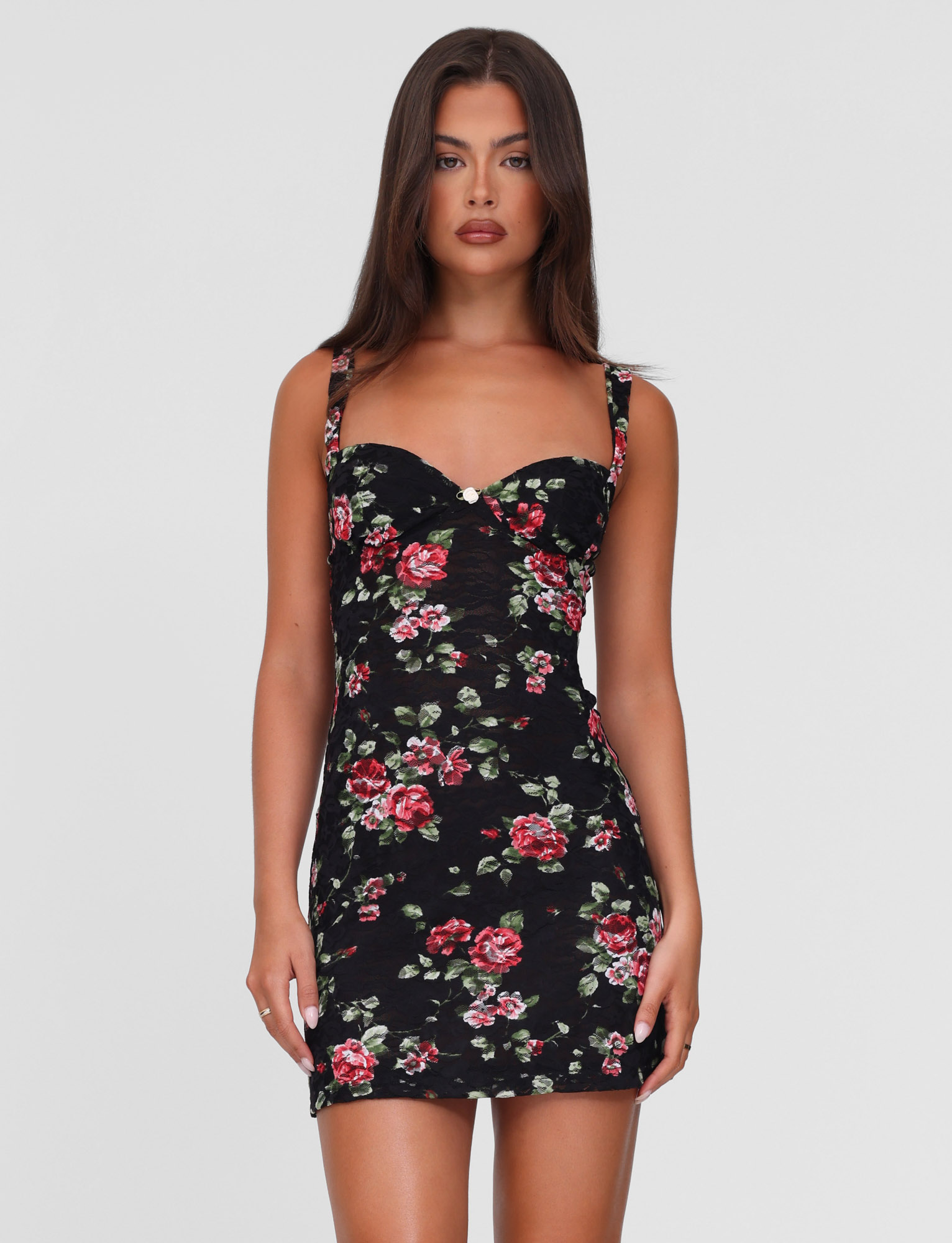 A woman with long brown hair wears the TIGERMIST ANDEE MINI DRESS in BlackRose floral, featuring a fitted, sleeveless lace design with red and pink rose prints, standing against a plain white background.