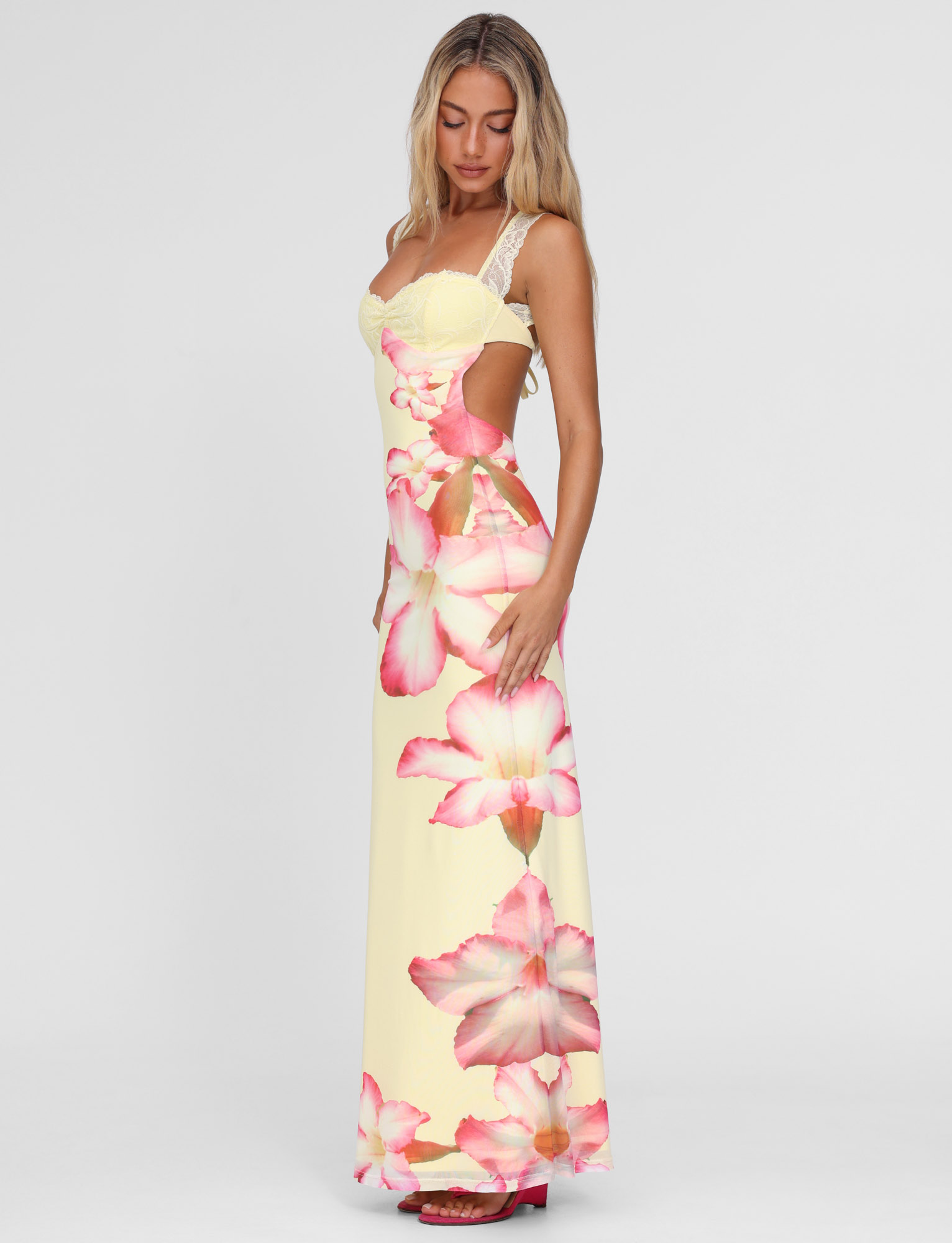 Woman with long blonde hair wearing the TIGERMIST NYOMI MAXI DRESS - YELLOW : MACRO FLORAL : YELLOW HIBISCUS, featuring lace straps, lace bust panel, and side cutouts, standing against a plain white background.