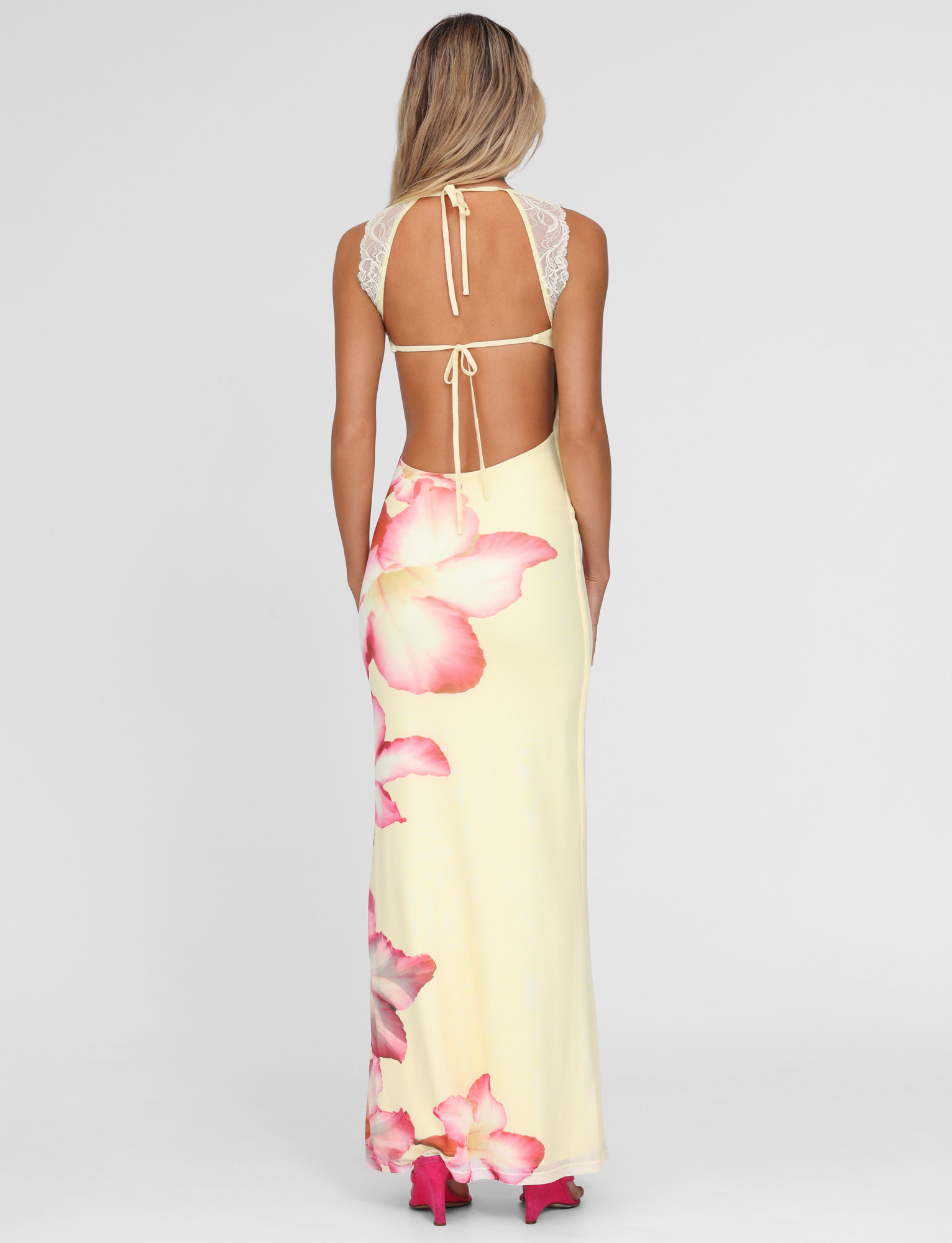 A woman with long blonde hair wears the TIGERMIST NYOMI MAXI DRESS in Yellow Hibiscus Macro Floral, featuring lace accents on the shoulders and bust, paired with pink heels against a plain white background.