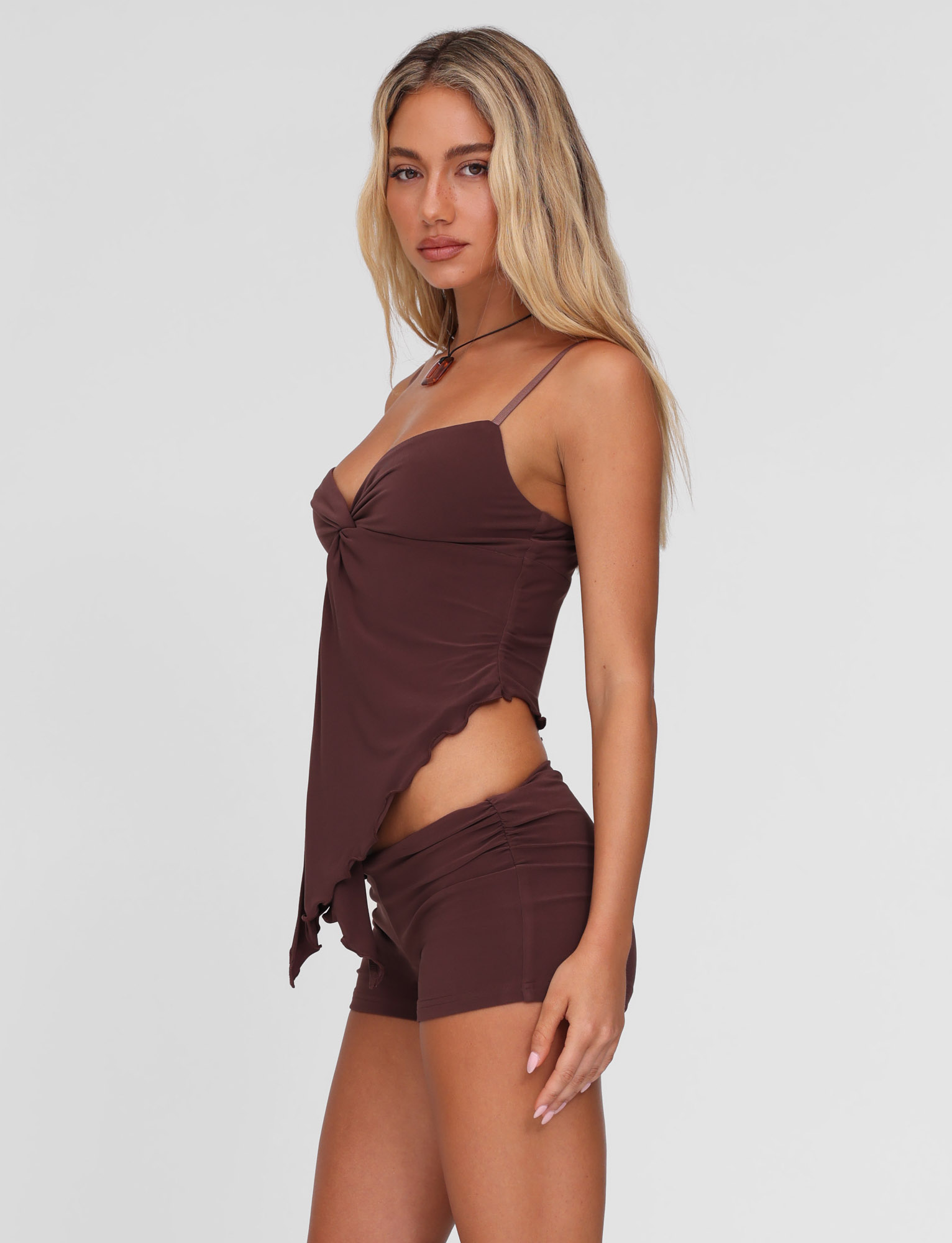 A woman with long blonde hair wears the TIGERMIST JOVIENNE TOP in chocolate brown, featuring adjustable straps, paired with matching brown shorts, standing against a plain light gray background.