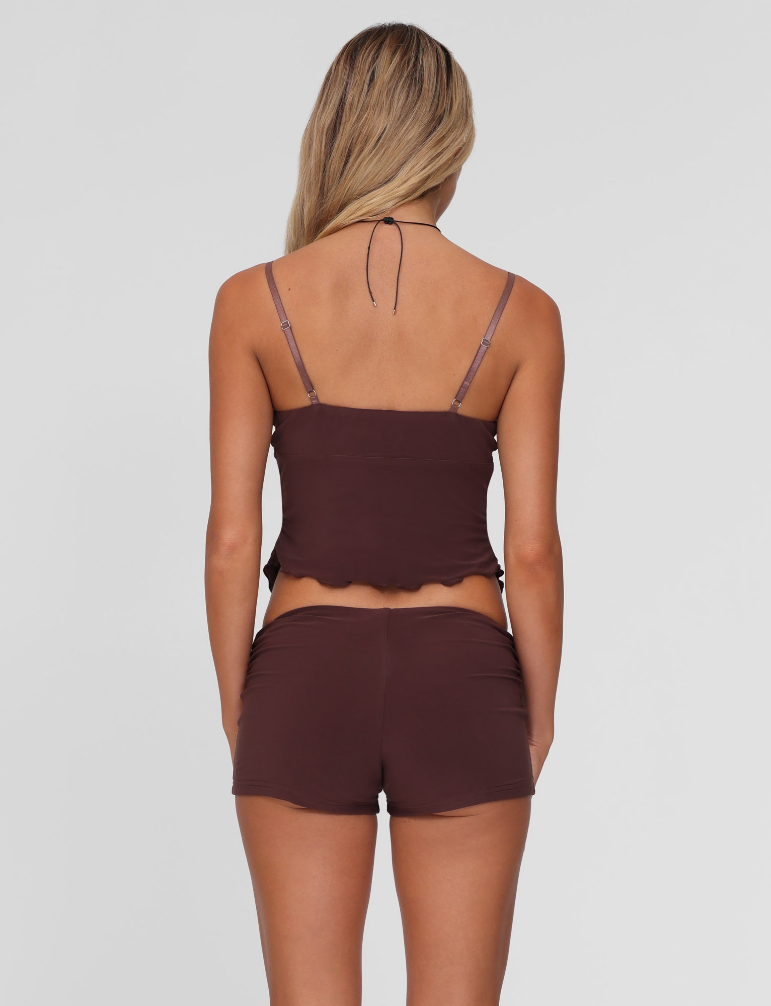 A woman with long, blonde hair is seen from the back wearing TIGERMIST’s JOVIENNE SHORT - BROWN : CHOCOLATE set, featuring a chocolate brown jersey camisole with thin straps and matching mid-rise shorts against a plain light background.