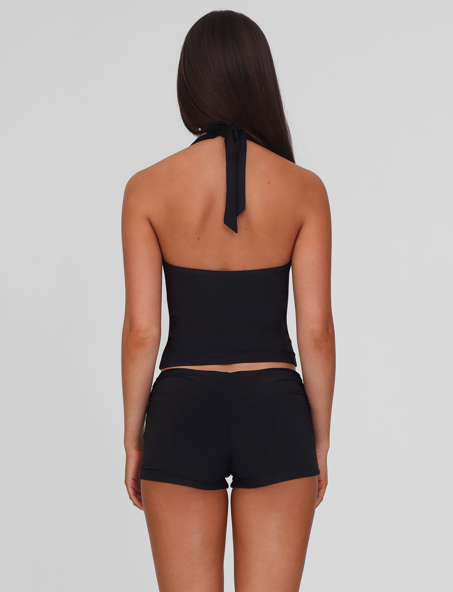 A woman with long brown hair stands with her back to the camera, wearing a black halter top and TIGERMIST JOVIENNE SHORT - BLACK jersey mini shorts with a fitted silhouette, set against a plain light gray background.