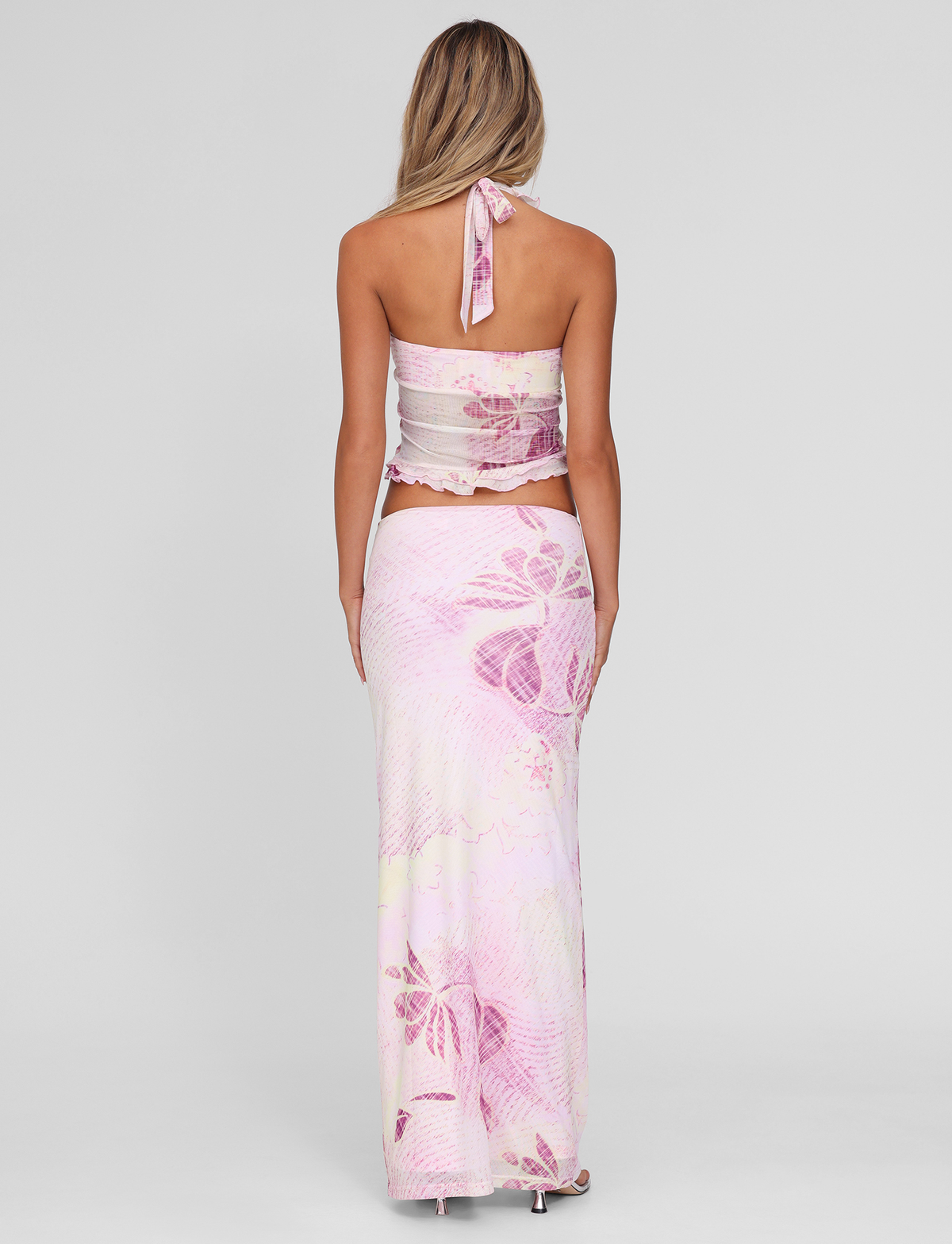 A woman with long blonde hair stands against a light gray background, wearing the TIGERMIST DALILA MIDI SKIRT in Grunge Floral Magenta with a matching halter top.
