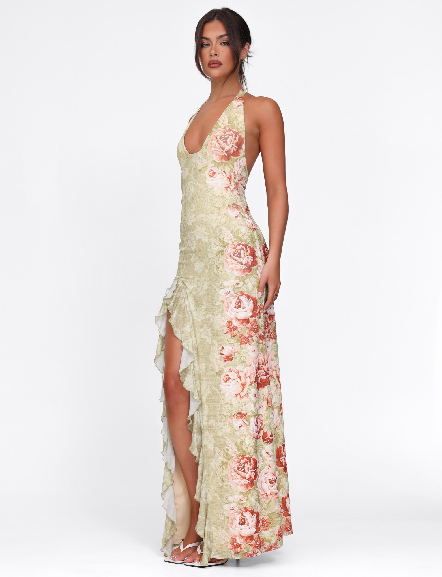 A woman models the TIGERMIST SIRENA MAXI DRESS in yellow with floral rose stripe print, featuring a halter neck and ruffle details. She pairs it with white heeled sandals and a loose updo against a plain white background.