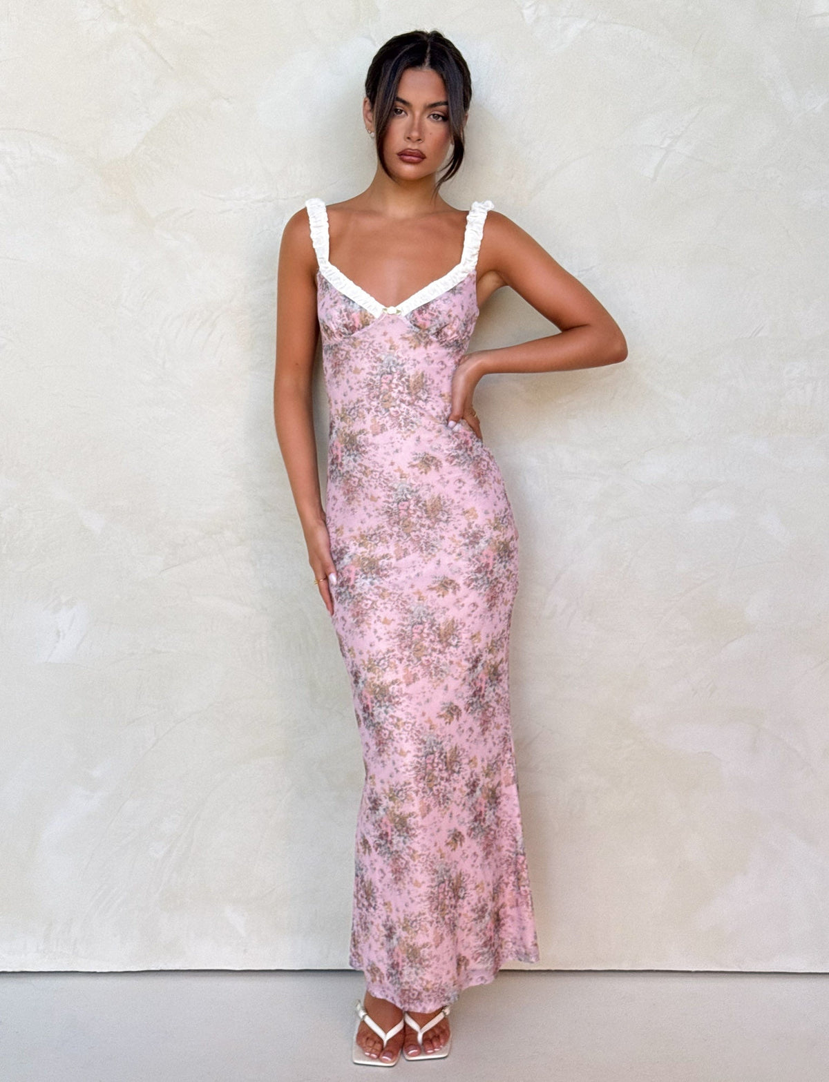 A woman with dark hair up poses confidently against a light textured wall, wearing the TIGERMIST AMARAN DRESS - PINK : CAMO FLORAL with white straps and neckline, paired with white heeled sandals.