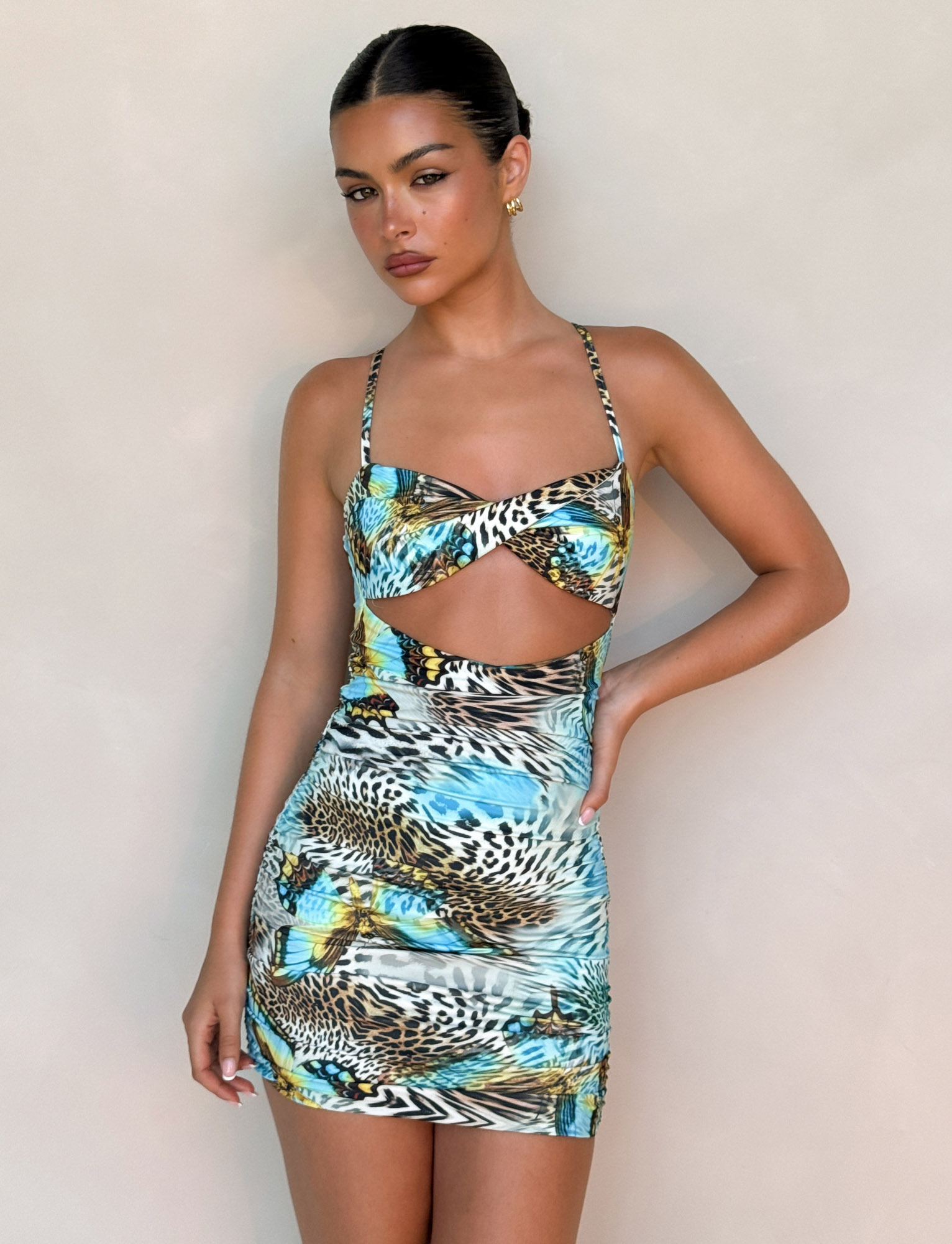 A woman with slicked-back hair stands against a plain background, confidently wearing the TIGERMIST VERA MINI DRESS - MULTI: ANIMAL: BUTTERFLY ANIMAL, featuring thin straps and a front cutout.