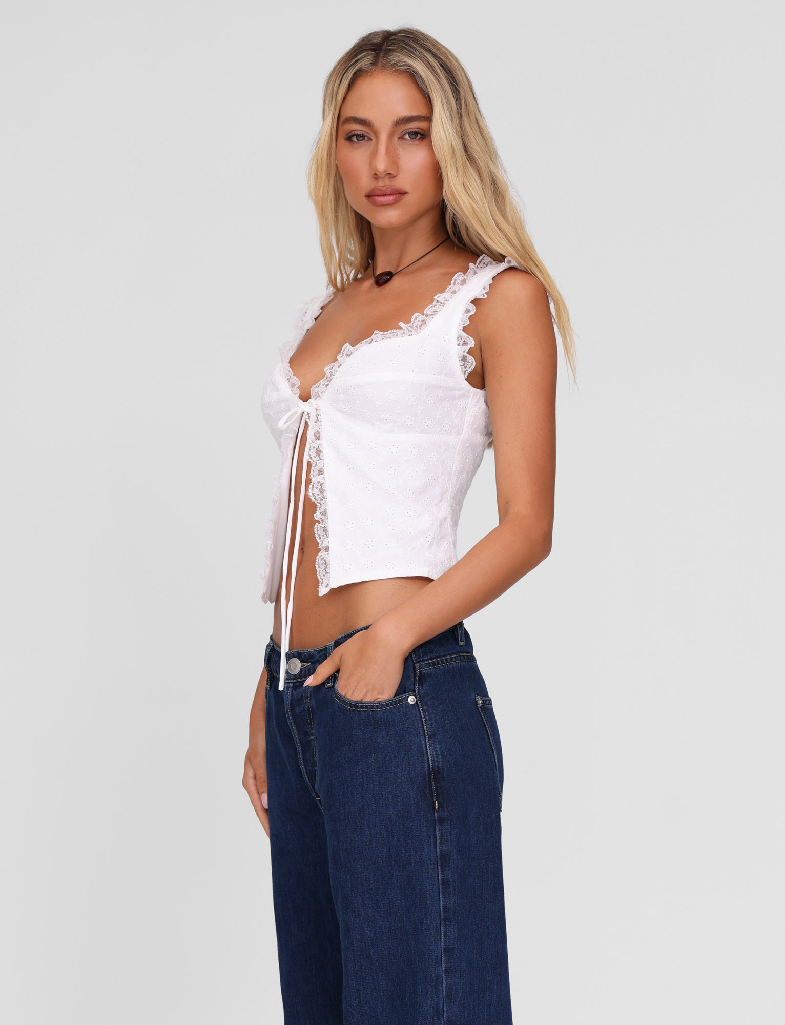 A woman with long blonde hair stands against a plain background, wearing the TIGERMIST KYANA TOP in white and blue jeans. She faces slightly sideways with one hand in her pocket, looking at the camera with a neutral expression.
