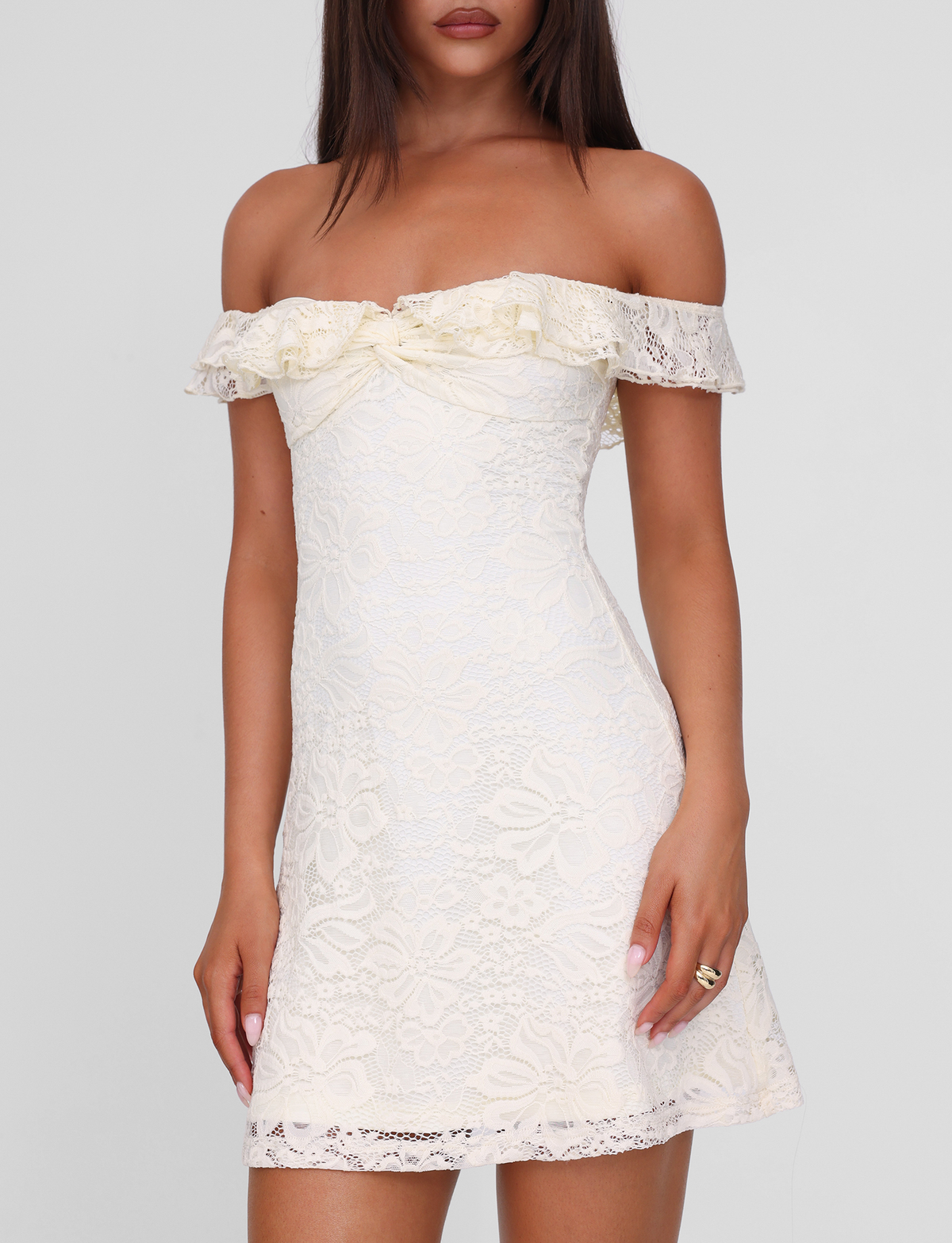 A woman wears the TIGERMIST DENALI LACE MINI DRESS in cream lace, featuring off-shoulder style and ruffled neckline, fitted through the body, standing against a plain light background.