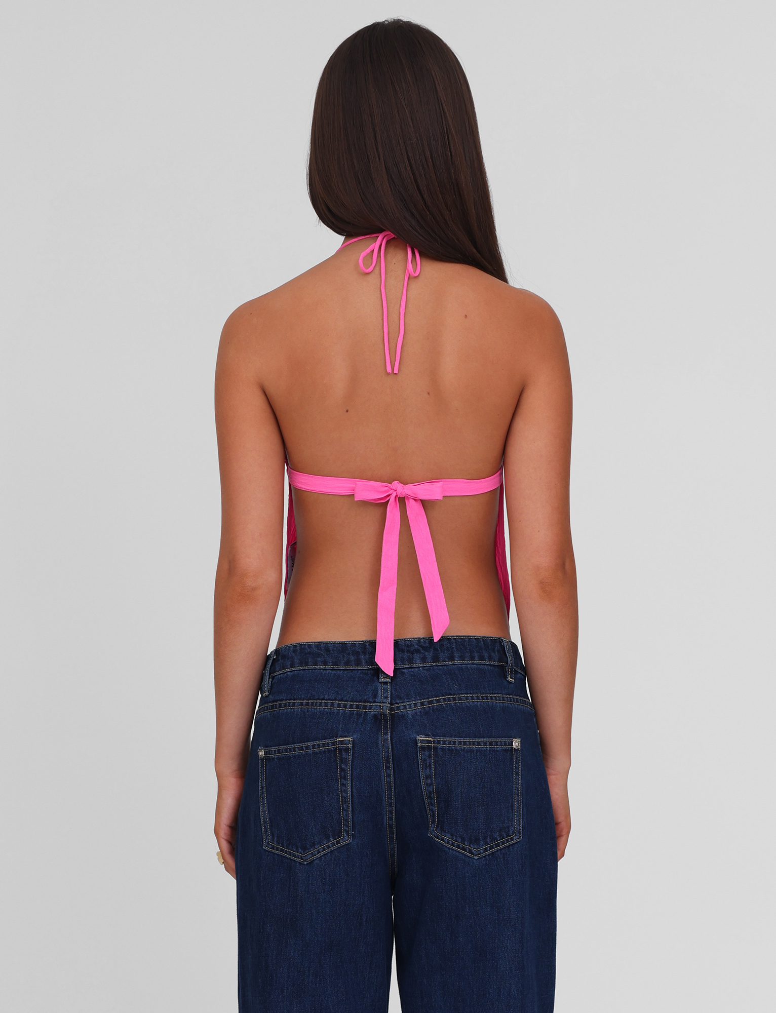 A person with long brown hair, seen from behind, models the TIGERMIST KAELAH TOP in hot pink, tied at the neck and back, paired with dark blue jeans featuring visible back pockets against a plain light gray background.